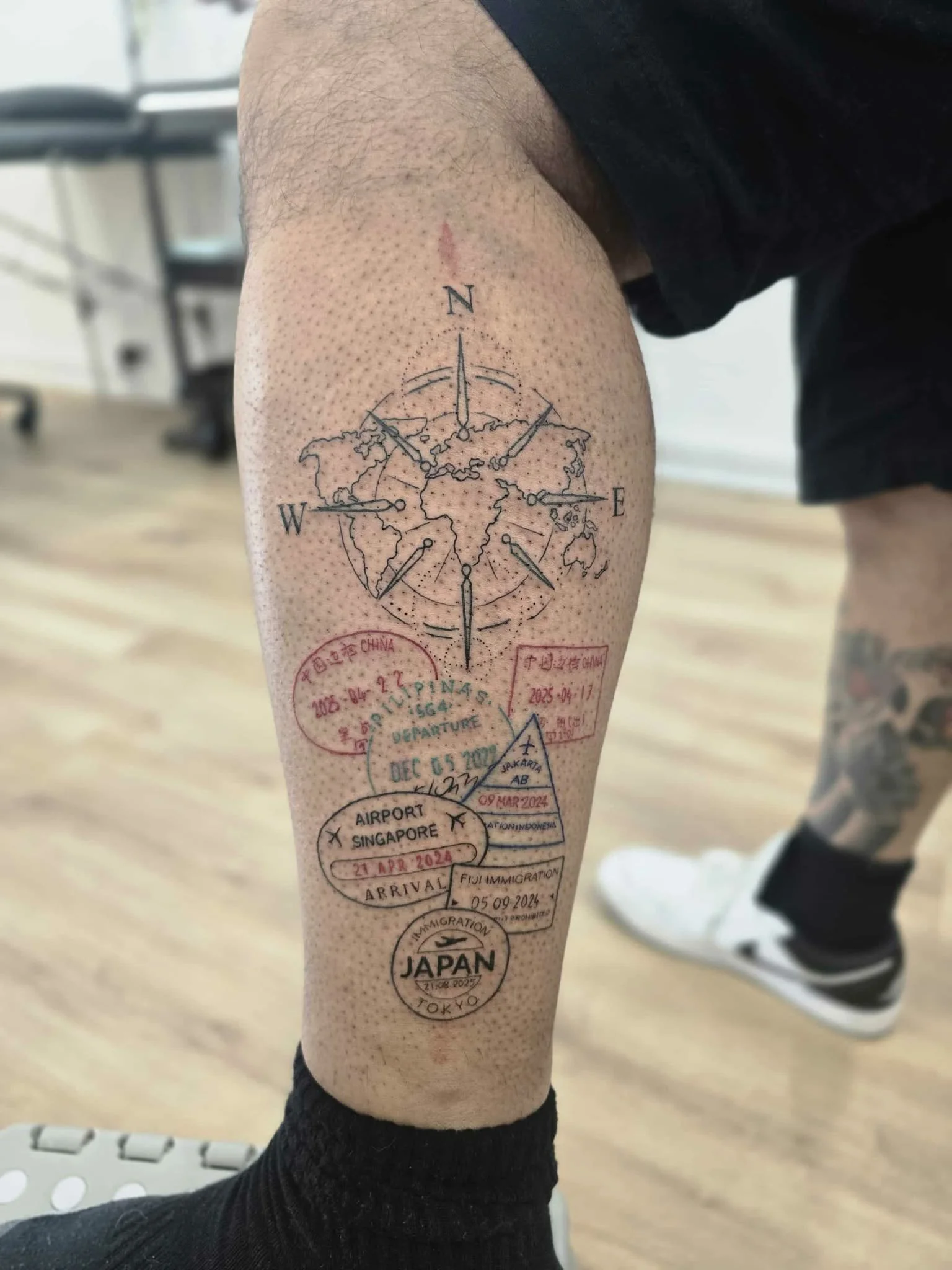 Michael baker tattoo. Tattoo on person's leg featuring a world map with a compass, airline stamps, and travel details including Singapore, Japan, and Indonesia. Fine line rose tattoo by Heart and Home Tattoo, Moonee Ponds Melbourne