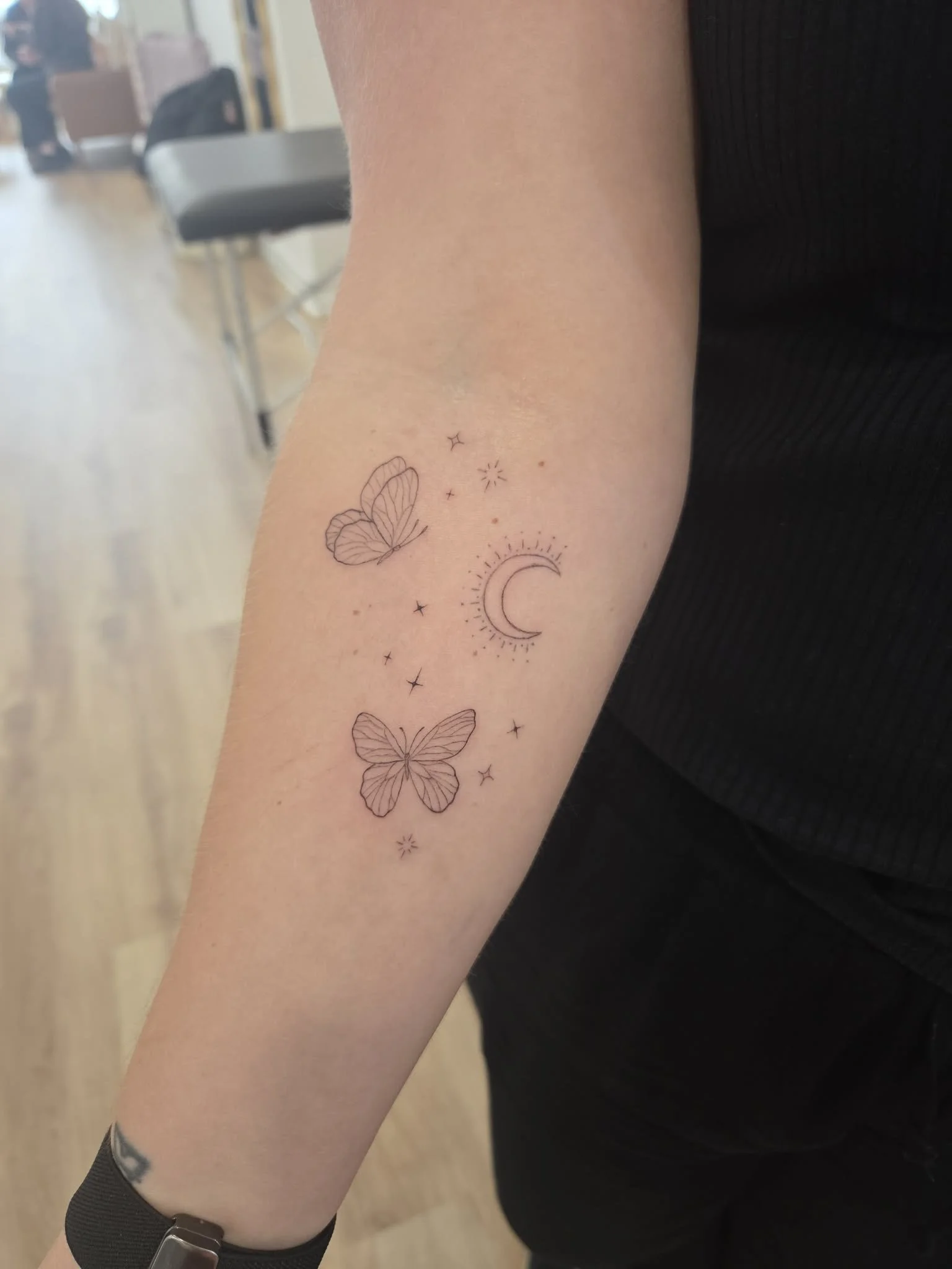 Fine line Tattoo of two outlined butterflies, a crescent moon with surrounding lines, stars, and small decorative dots on a person's forearm.