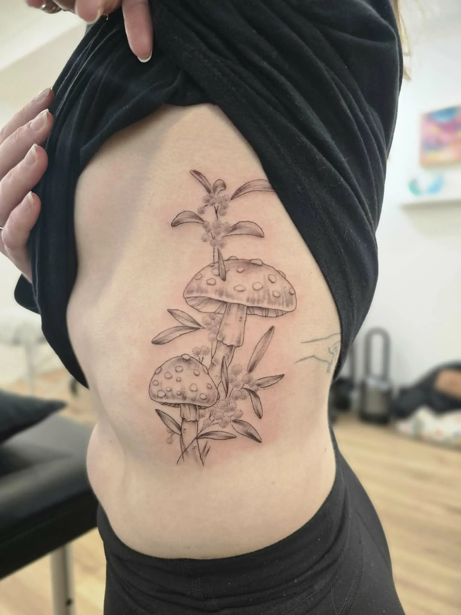 A woman is showing her side tattoo of mushrooms with leaves, done in black ink with fine lines.