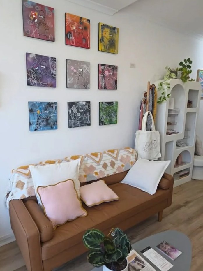 Living room corner with a brown sofa decorated with pink and white pillows, a white blanket with orange patterns, a round coffee table with magazines, a potted plant, and colorful abstract artwork on the wall. There is a white shelving unit with books and plants next to the sofa.