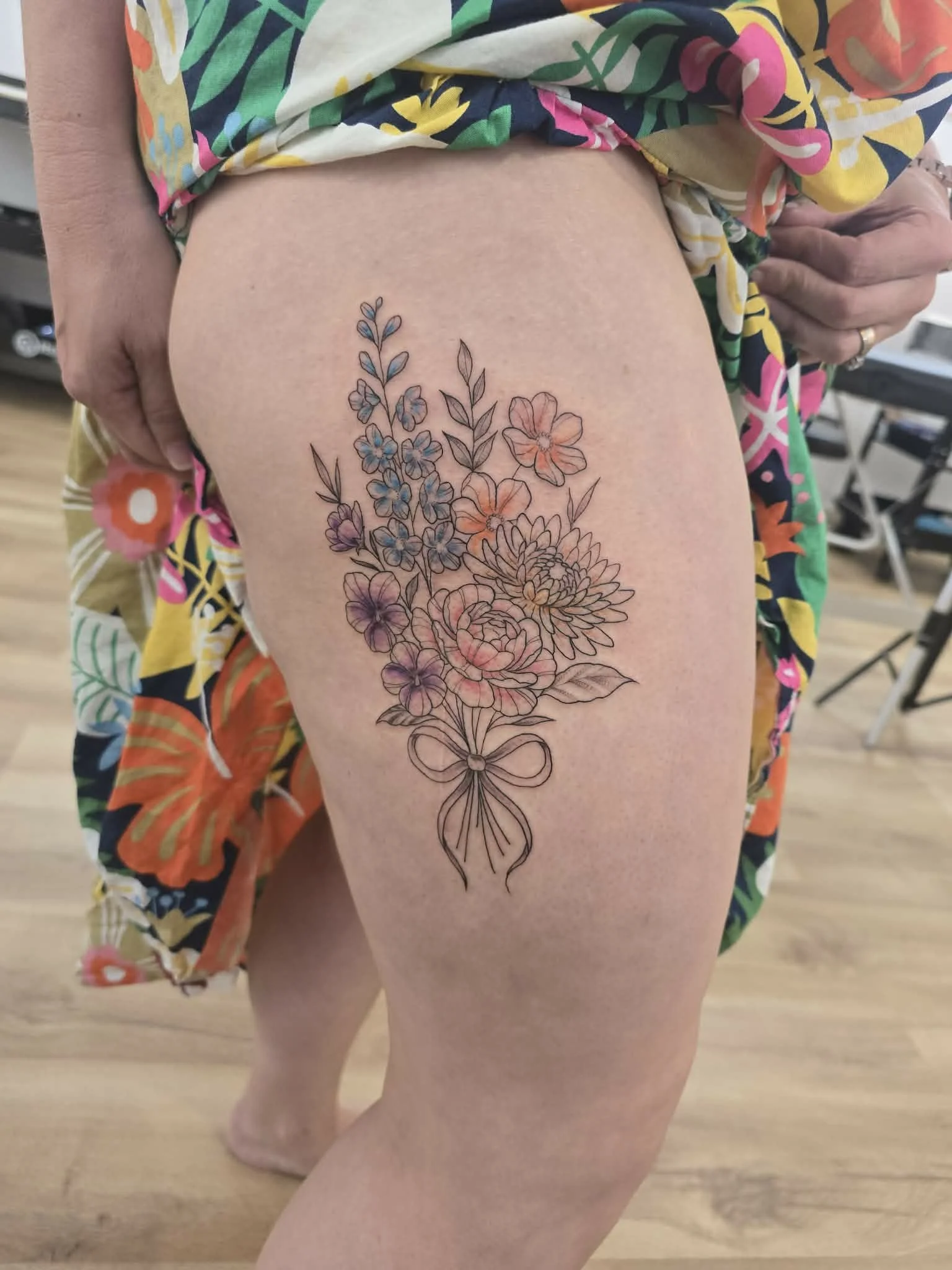 A floral tattoo featuring various flowers and leaves, outlined with some colored shading, on a person's thigh.