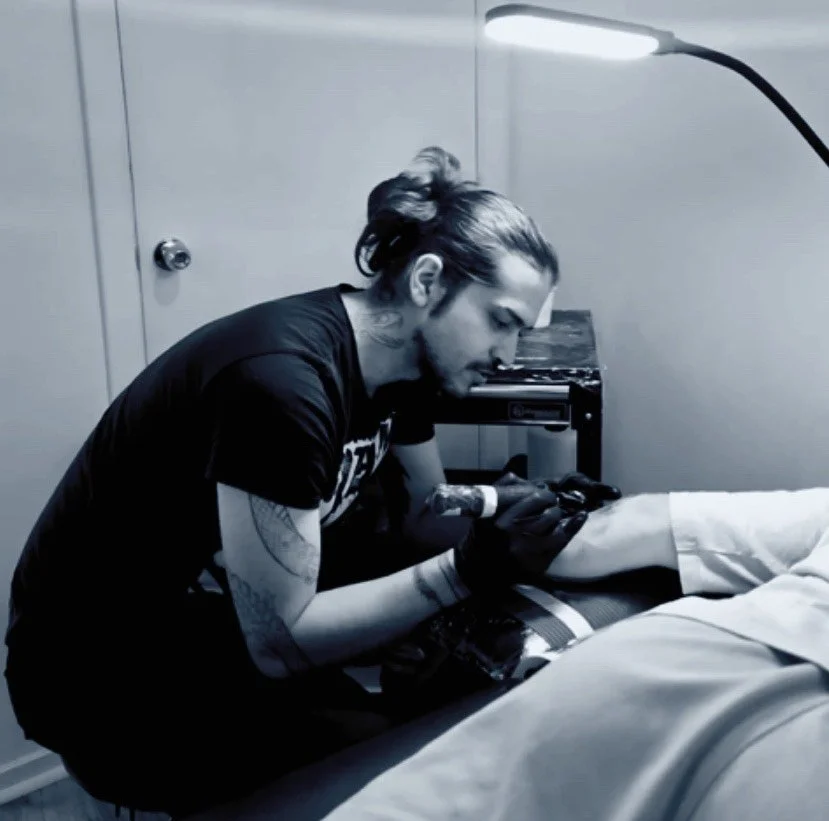 A tattoo artist working on a client's arm in a monochrome room, sitting at a table with tattoo equipment.