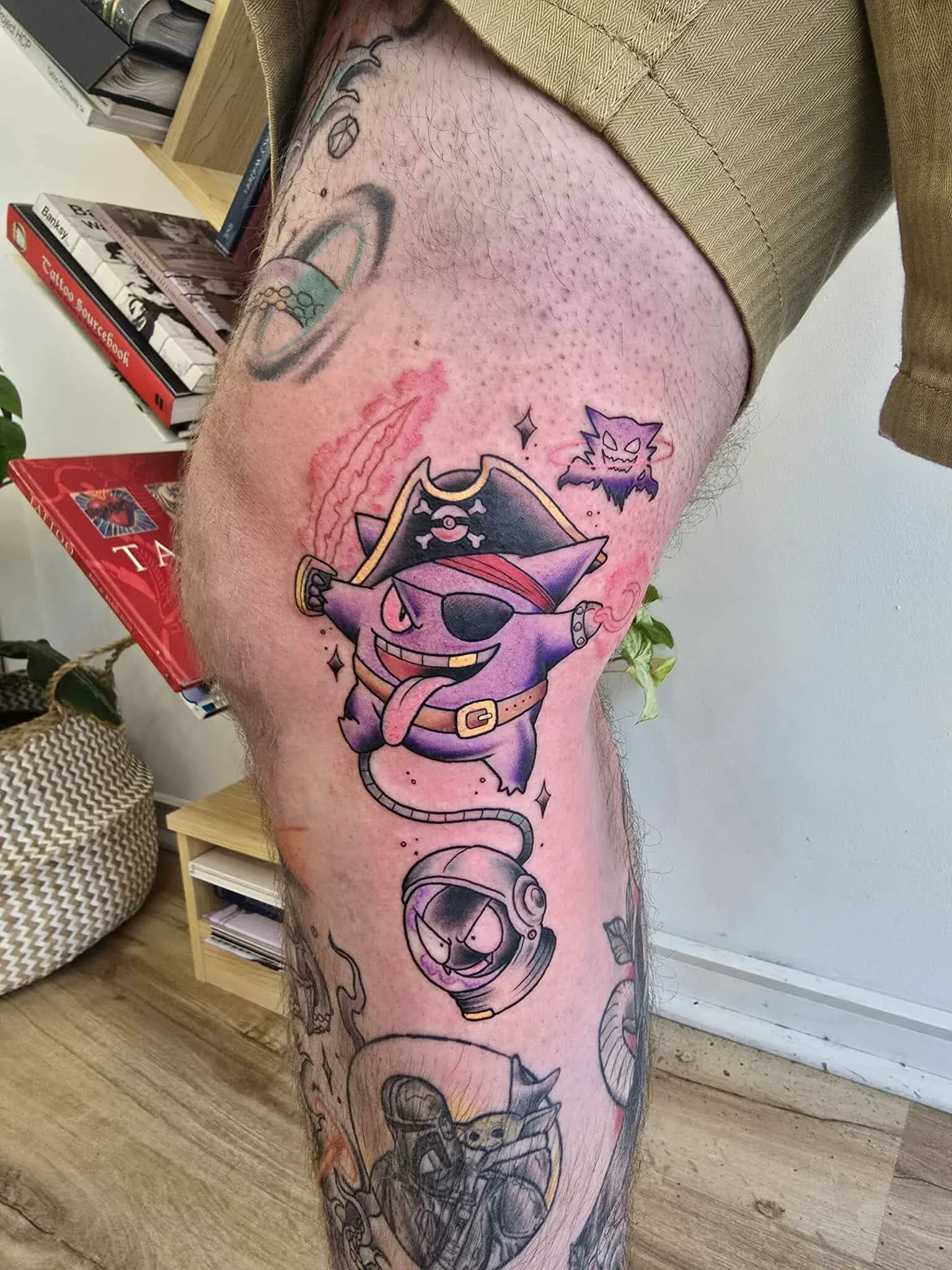 Colorful tattoo of a cartoon pirate cat sticking out its tongue, wearing an eye patch and pirate hat, with a pirate hook on one paw, connected by a line to a space helmet with a cat face inside, on a person's thigh.
