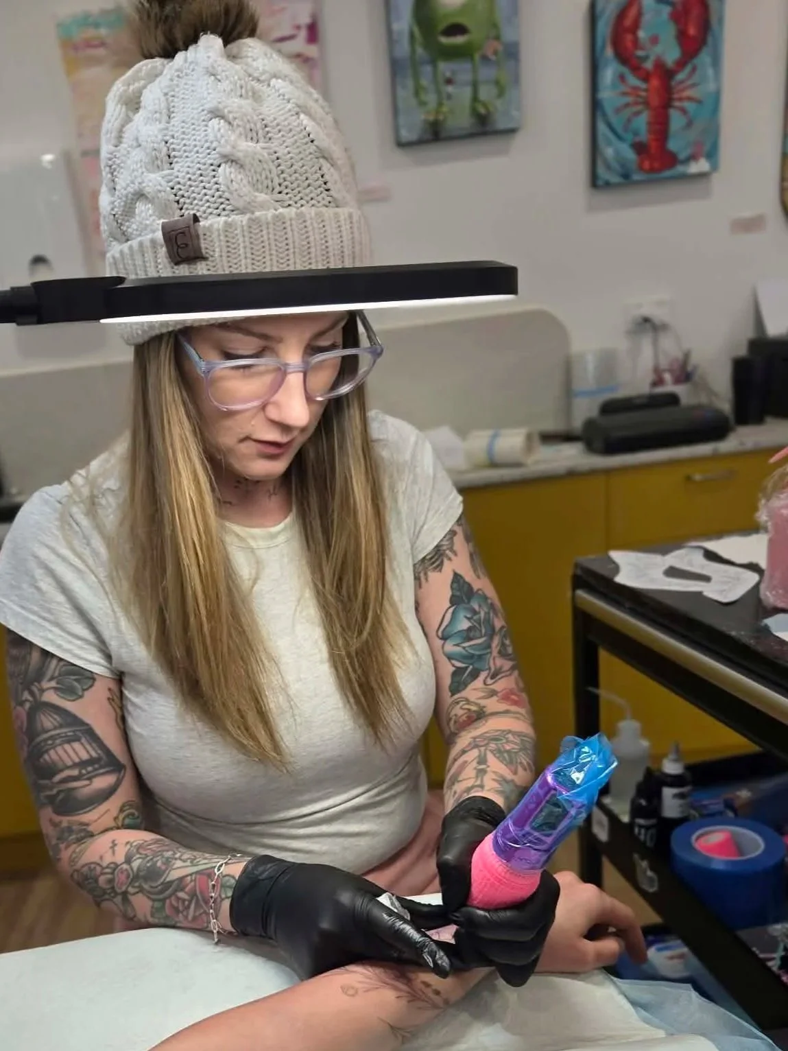 Tattooed woman wearing a beige shirt, glasses, and a knit hat getting a tattoo on her forearm in a tattoo studio. Renee Baker Melbourne based tattoo artist, friendly tattoo artist in Melbourne, Female tattoo artist.