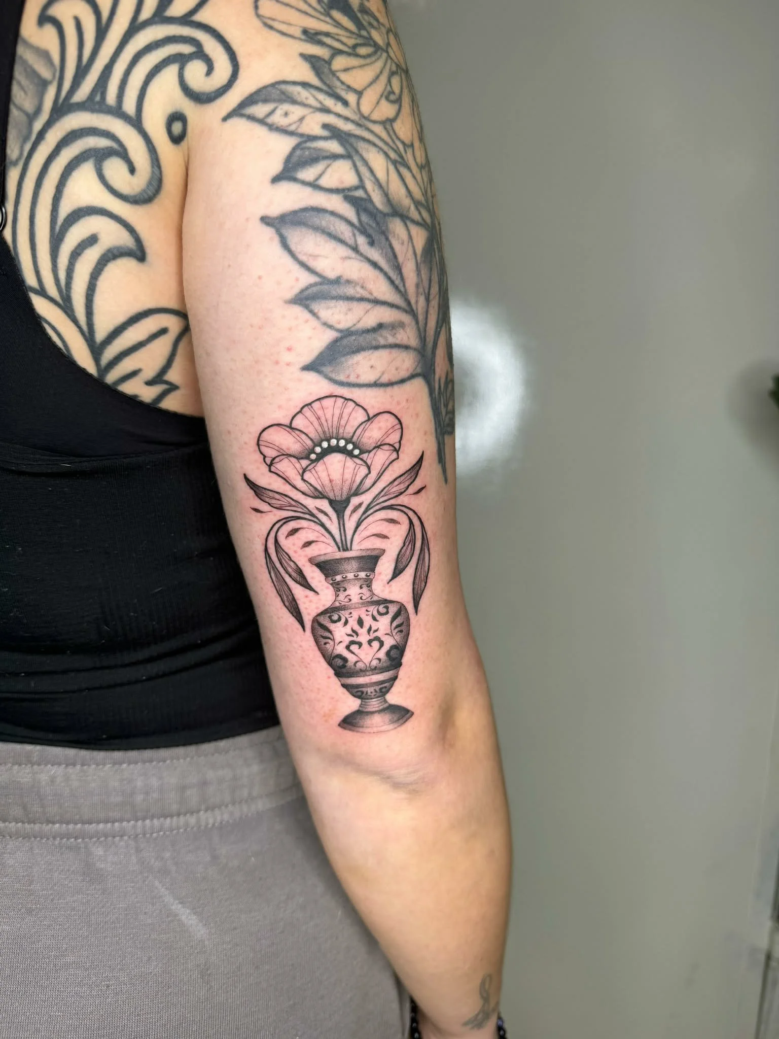 Tattoo of a flower in a decorative vase on a person's arm, with additional tattooed designs on the shoulder and upper arm.