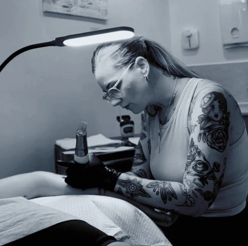 A tattooed woman wearing glasses and gloves, working on a client's arm in a tattoo studio, with a lamp overhead illuminating her work area.