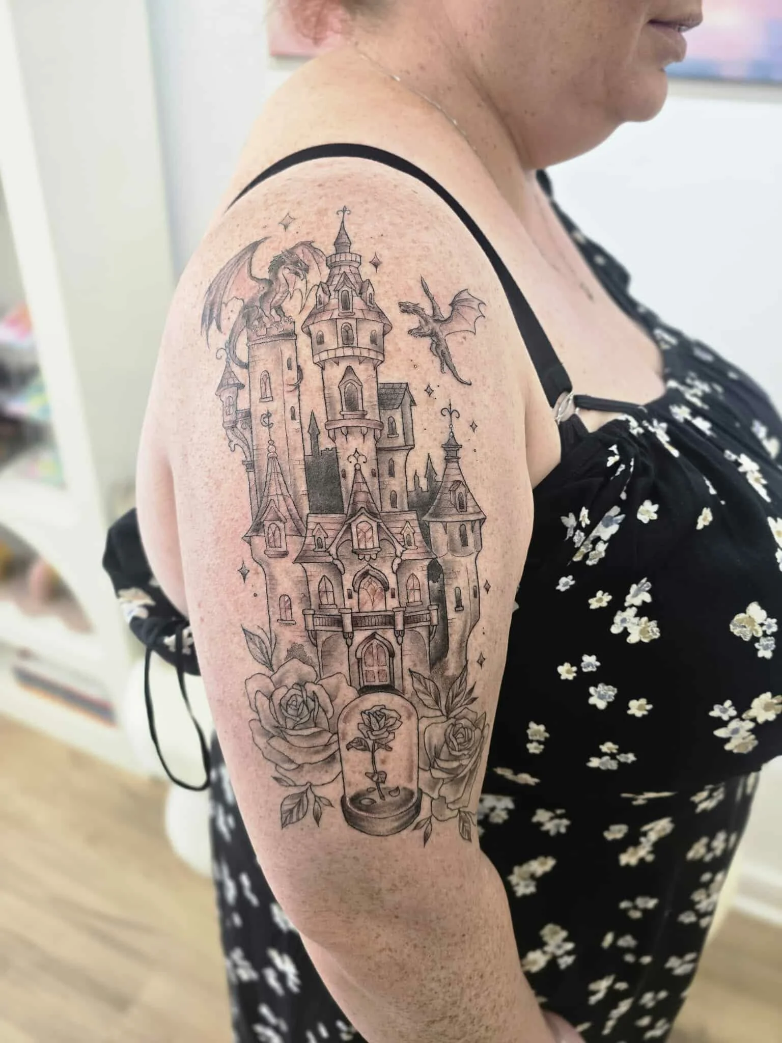 Fine Line Tattoo of a whimsical castle with towers, a dragon, and a rose with a snail shell at the bottom on a woman's shoulder. Detailed tattoo illustrative tattoo style