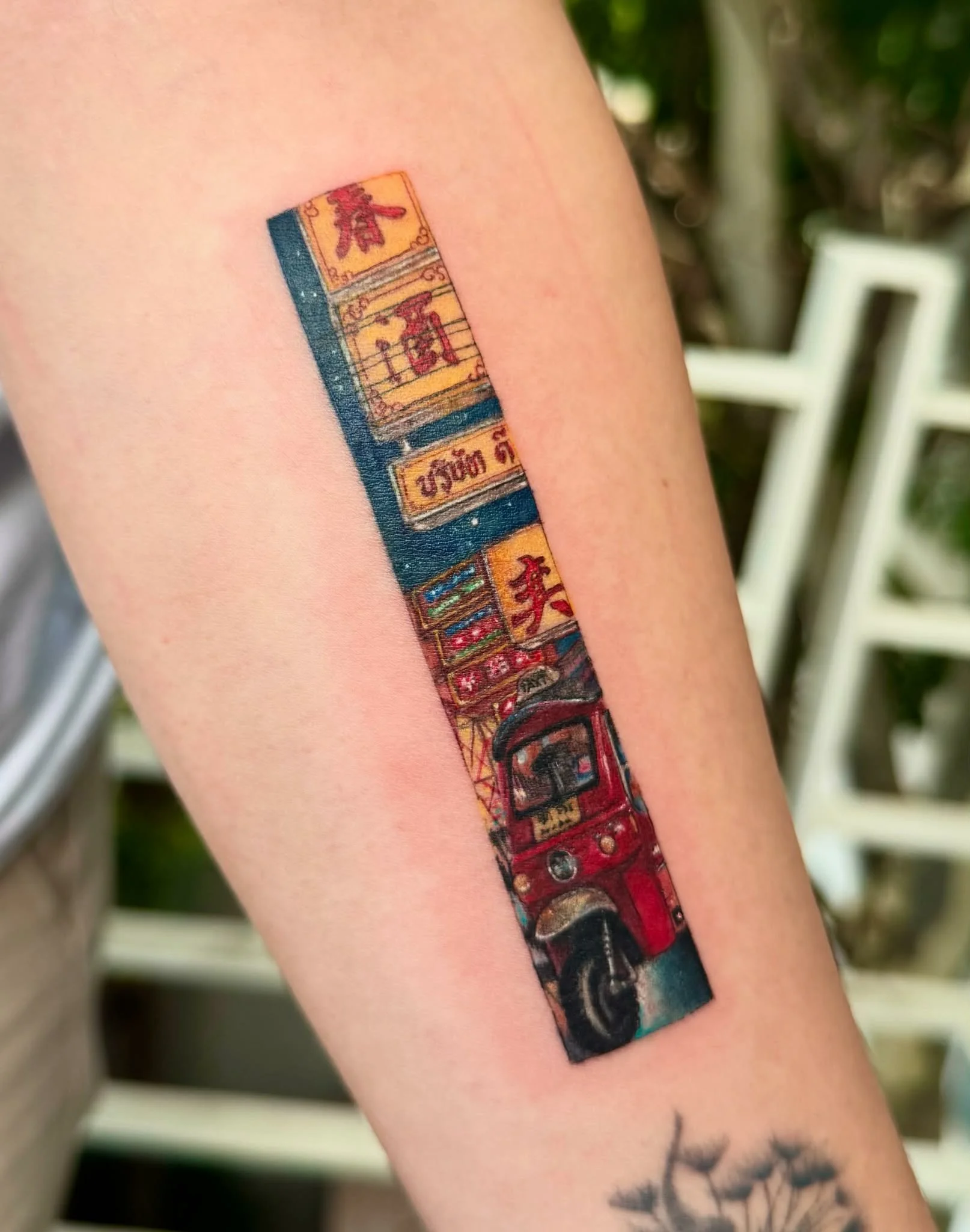 Tattoo of a red tuk-tuk with a city street scene, including signs with Thai script, on a person's arm.