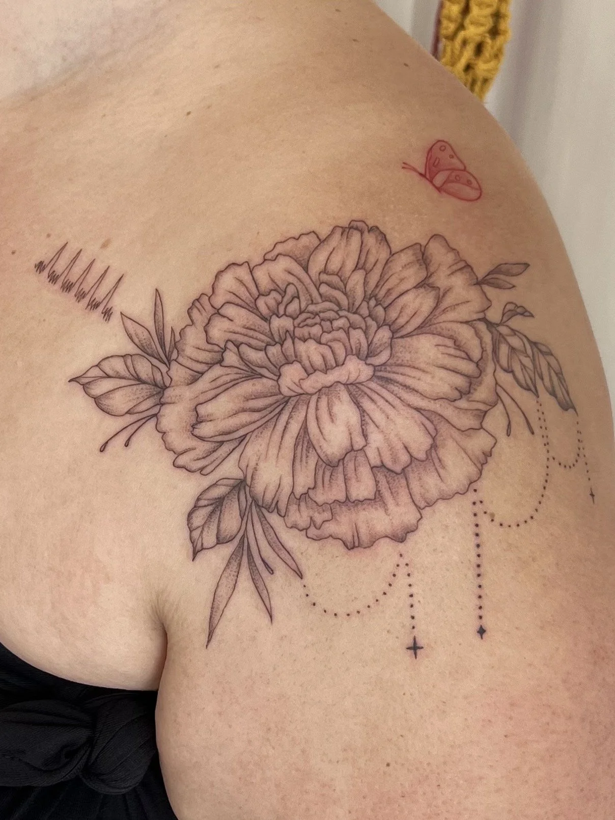 A detailed black ink tattoo of a large peony flower on a person's shoulder, with leaves and decorative dotted lines hanging beneath it, along with a small red ladybug tattoo near the collarbone.