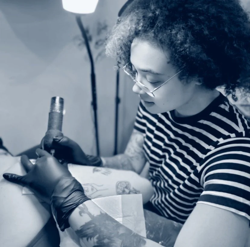 Tattoo artist in striped shirt with glasses creating tattoo on client's arm, tattoo machine in hand, working in studio with lamp overhead.