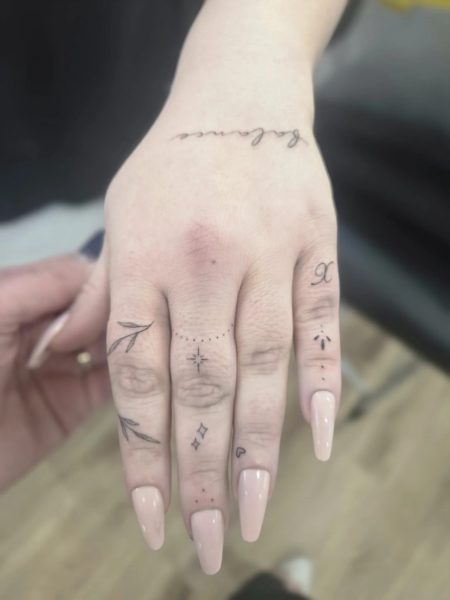 A hand with various small tattoos, including stars, dots, a heart, a leaf, and cursive text on the wrist.