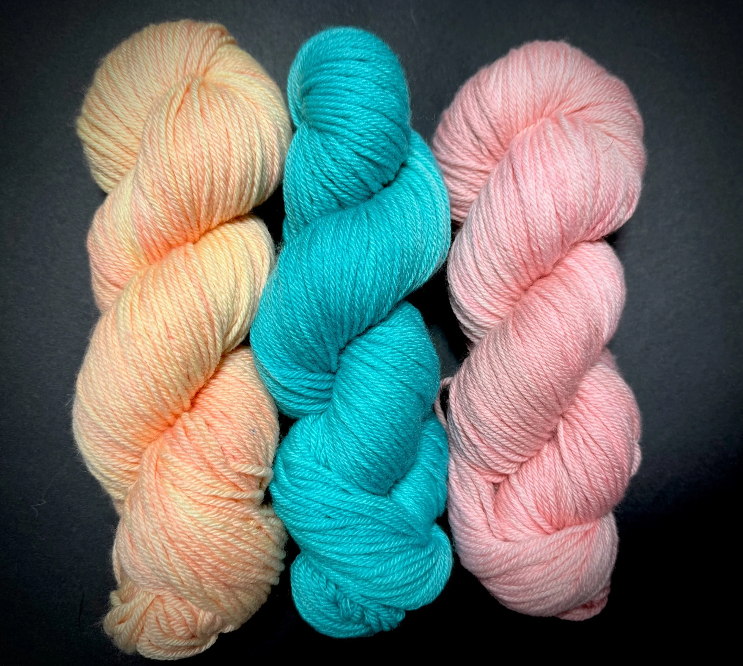 Three skeins of yarn in pastel colors: peach, turquoise, and pink, arranged on a black background.