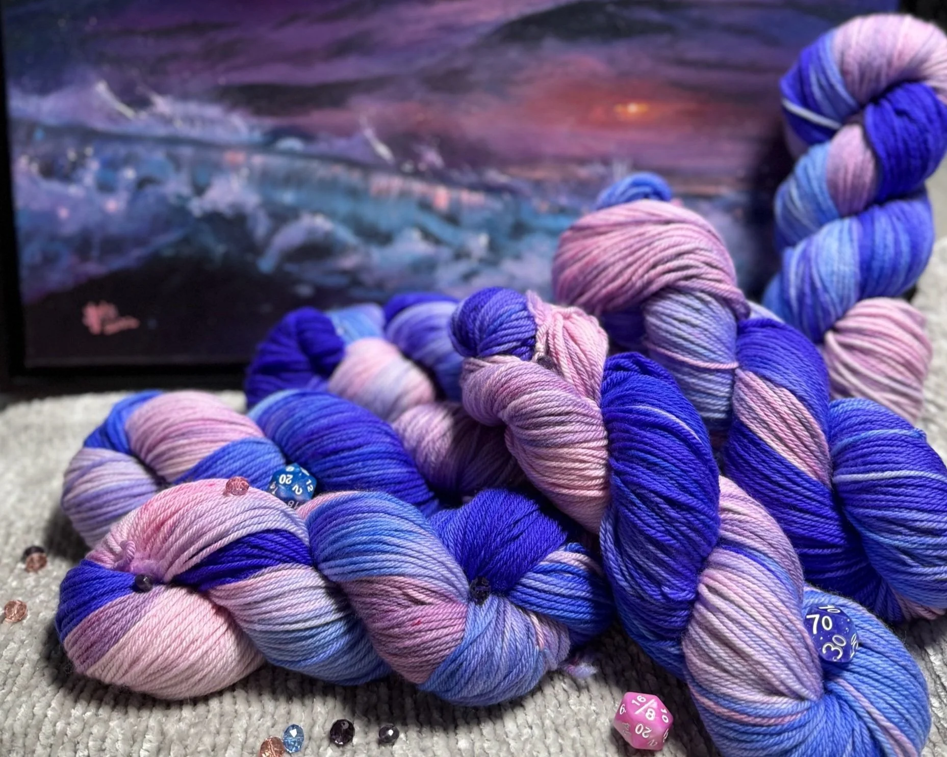 Colorful skeins of pink and blue variegated yarn with game dice and a painting in the background.