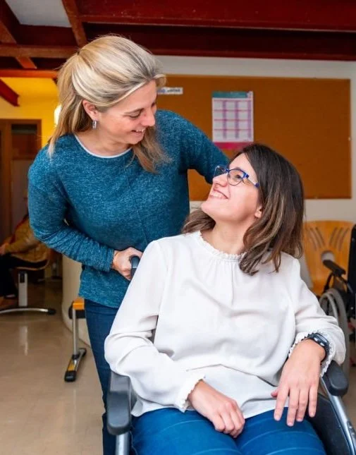 A woman with glasses sitting in a wheelchair smiling at another woman who is leaning over and smiling back at her in a warm, well-lit room.