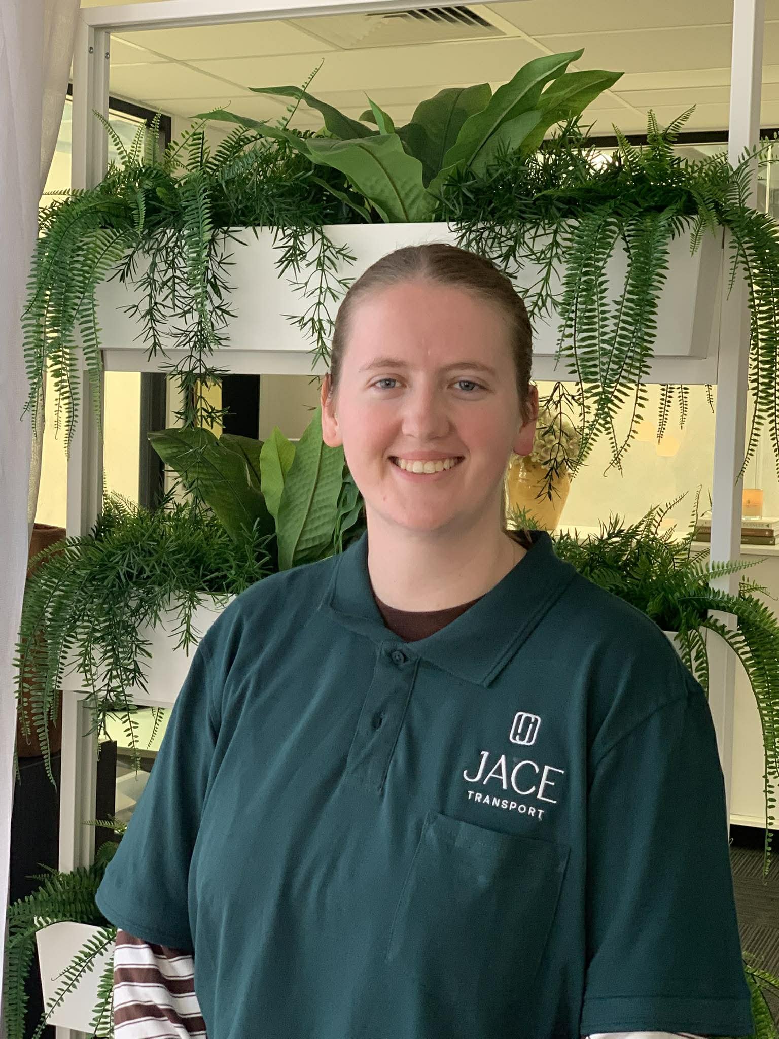 A smiling young man with light skin and brown hair, wearing a dark green shirt with a company logo and the words 'JACE TRANSPORT.' He is standing in front of a large white shelf filled with lush green indoor plants.