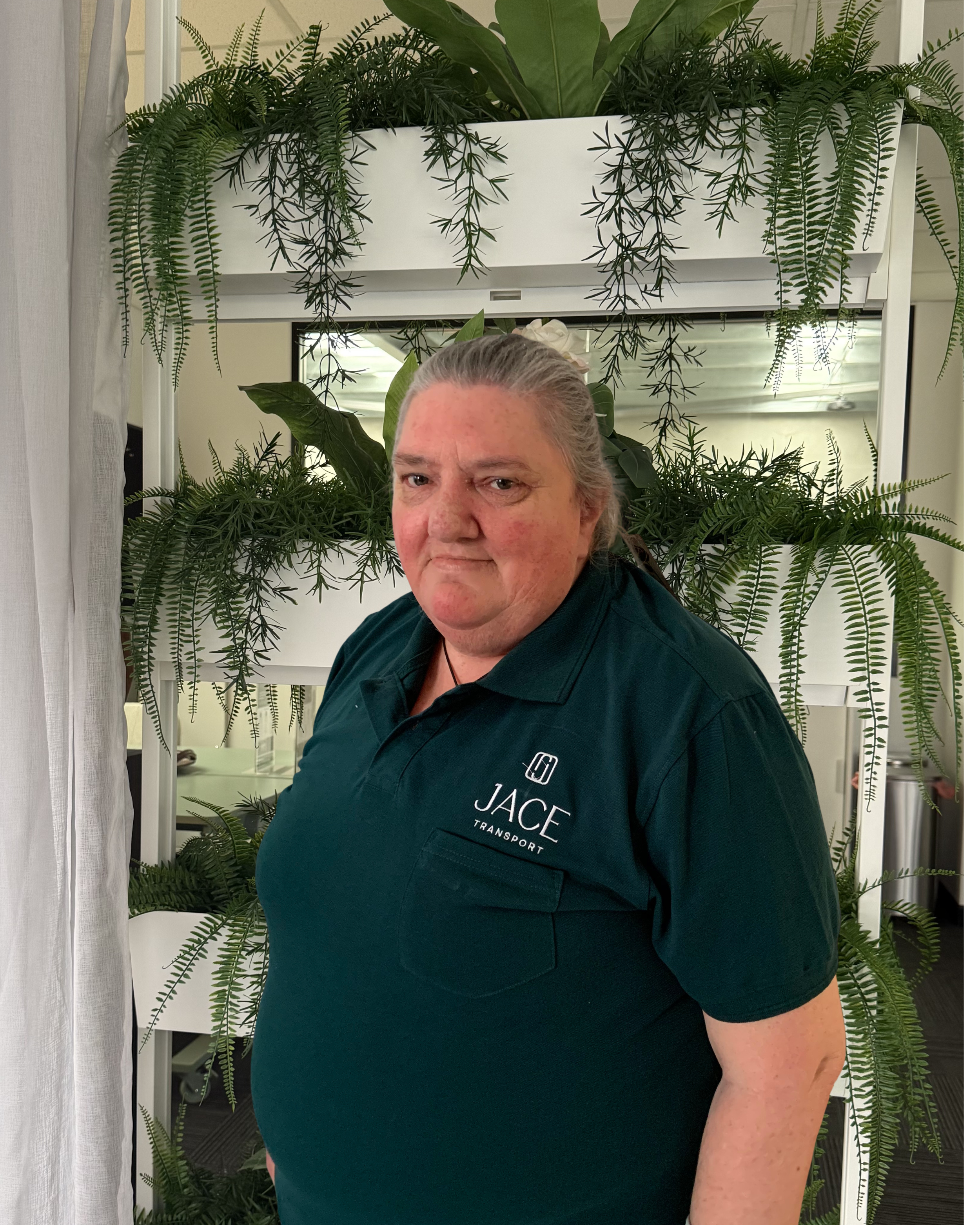 A middle-aged woman with grey hair tied back and wearing a dark green shirt with a company logo for JACE Transport, standing in front of a backdrop of green plants and a mirror.