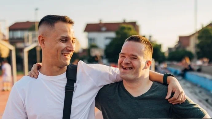 Two men smiling and hugging each other outdoors in a neighborhood or park.