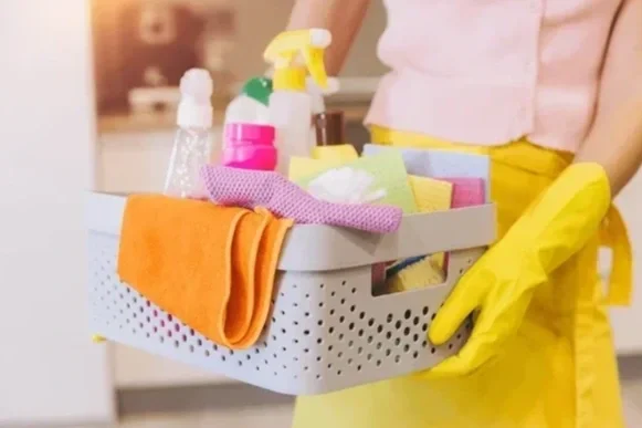 Person wearing yellow gloves holding a basket filled with cleaning supplies and cloths in a kitchen.