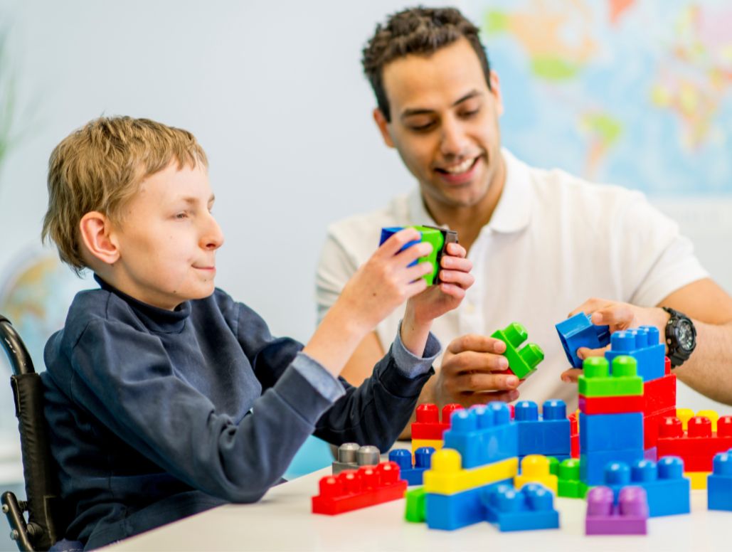 A young boy with a disability sitting at a table playing with colorful building blocks, with an adult male assisting him, in a bright room with a world map in the background.