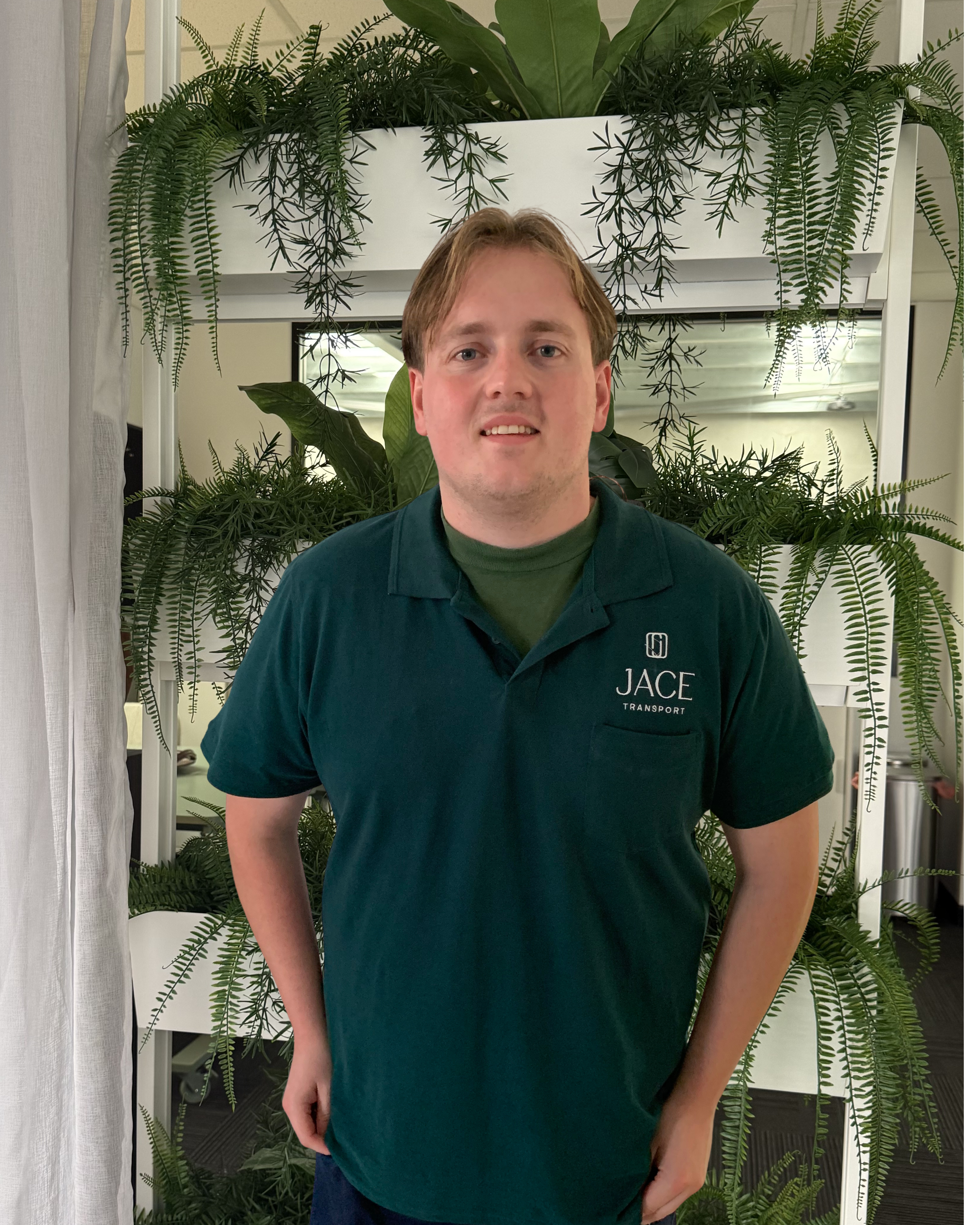 A man standing indoors in front of green plants and a mirror, wearing a dark green shirt with the logo 'JACE Transport'.
