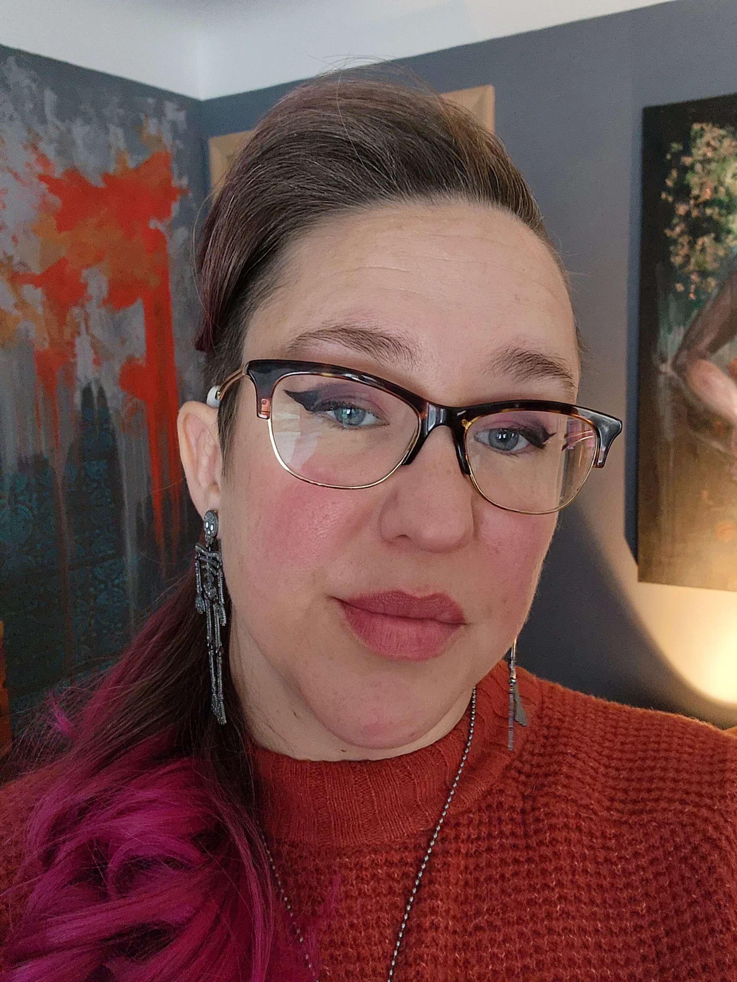 The artist seen from the shoulders up, wearing an orange sweater with pink hair, dark hornrimed glasses, and skeleton earrings.