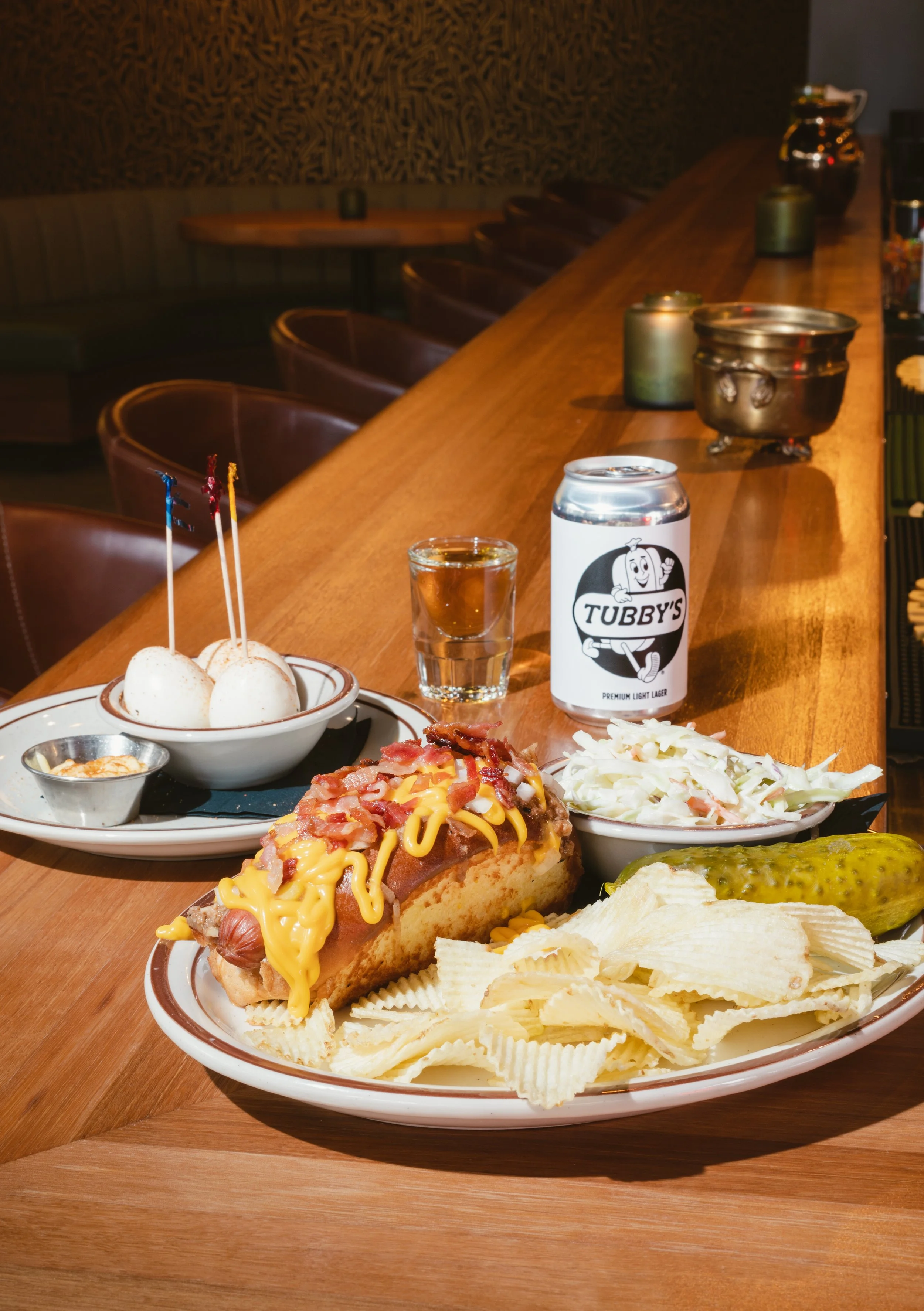 Food and drink on a counter at Tubby Bar in Calgary, including potato chips, relish, coleslaw, hot dog with cheese and bacon, pickle, a glass of water, a can of Tubby's beer, and a plate with eggs, with a bar seating background.