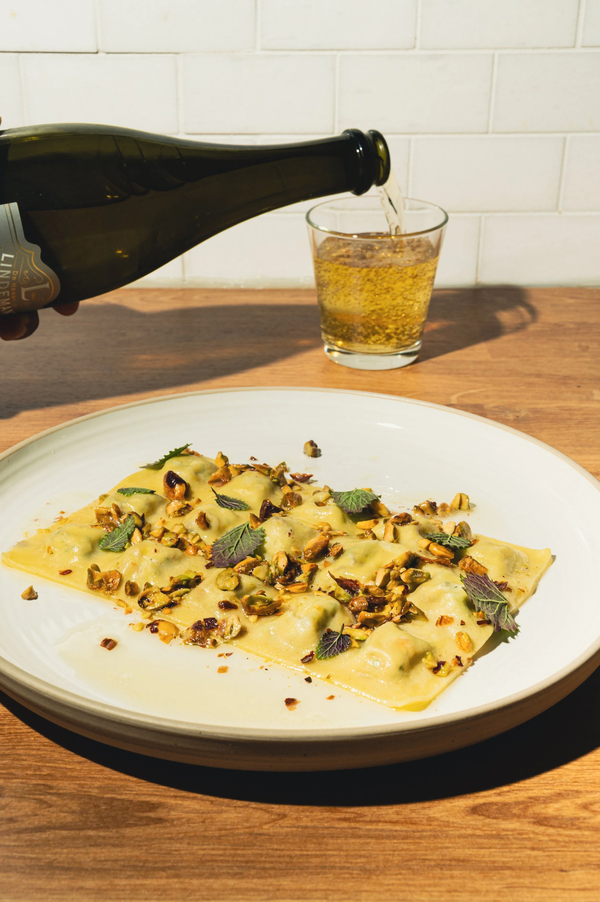 A white plate with ravioli topped with chopped pistachios and garnished with purple and green leaves, on a wooden table with a glass of beer and someone pouring beer from a bottle in the background.