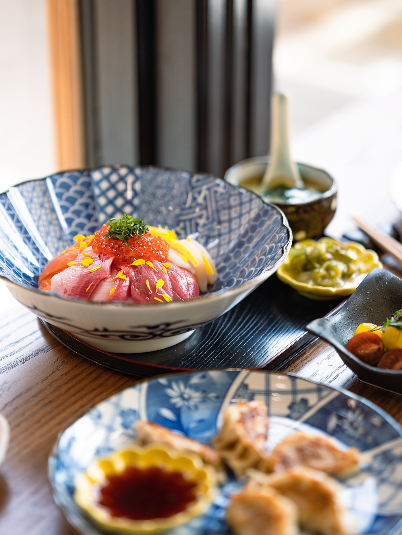 A bowl of sashimi with slices of raw fish topped with fish roe and garnished with herbs, surrounded by other small dishes of food, on a wooden table near a window at Satsuki in Calgary