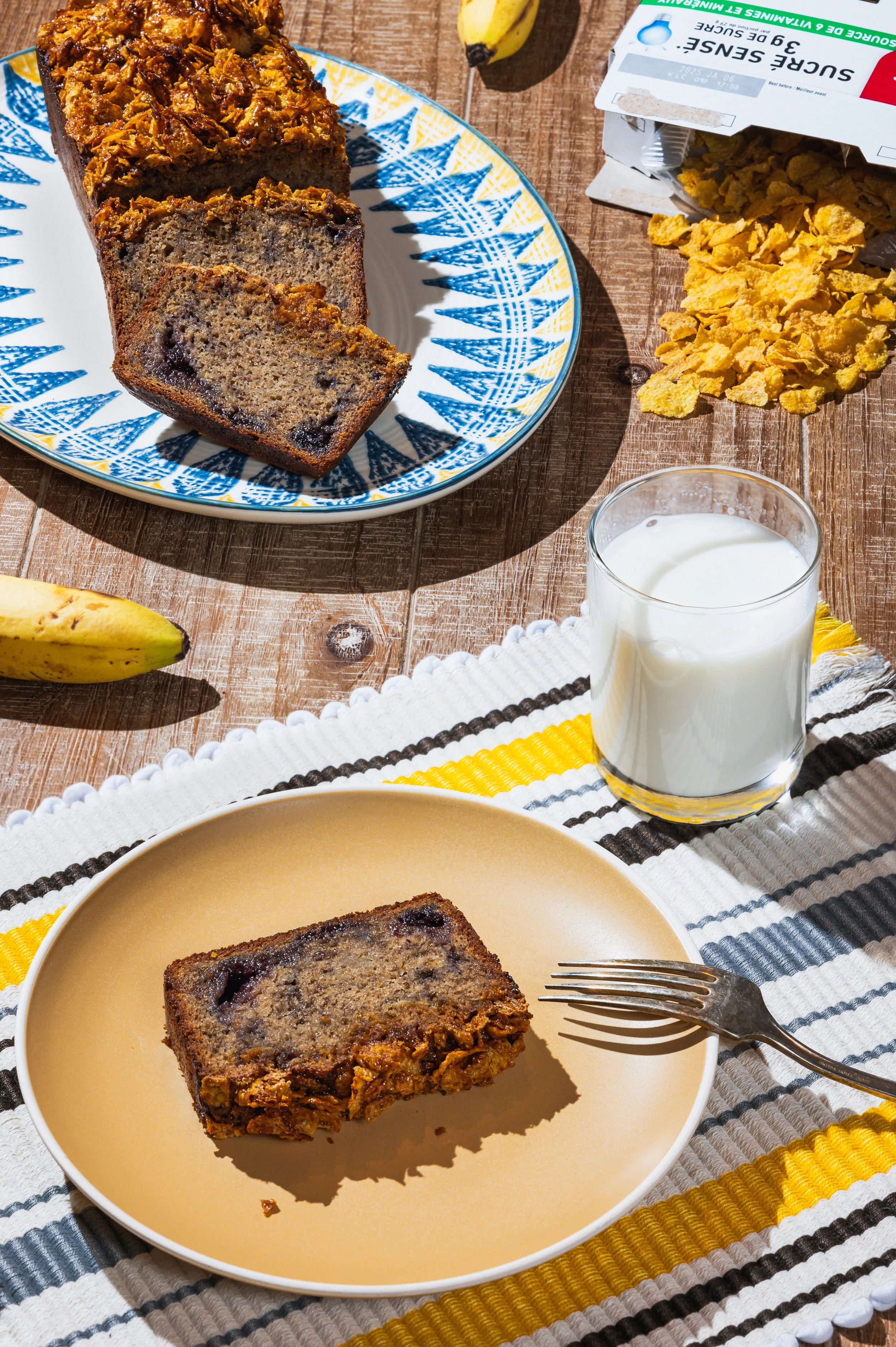 A slice of banana bread on a yellow plate with a fork, a glass of milk, bananas, cornflakes, and a baking box on a wooden table.