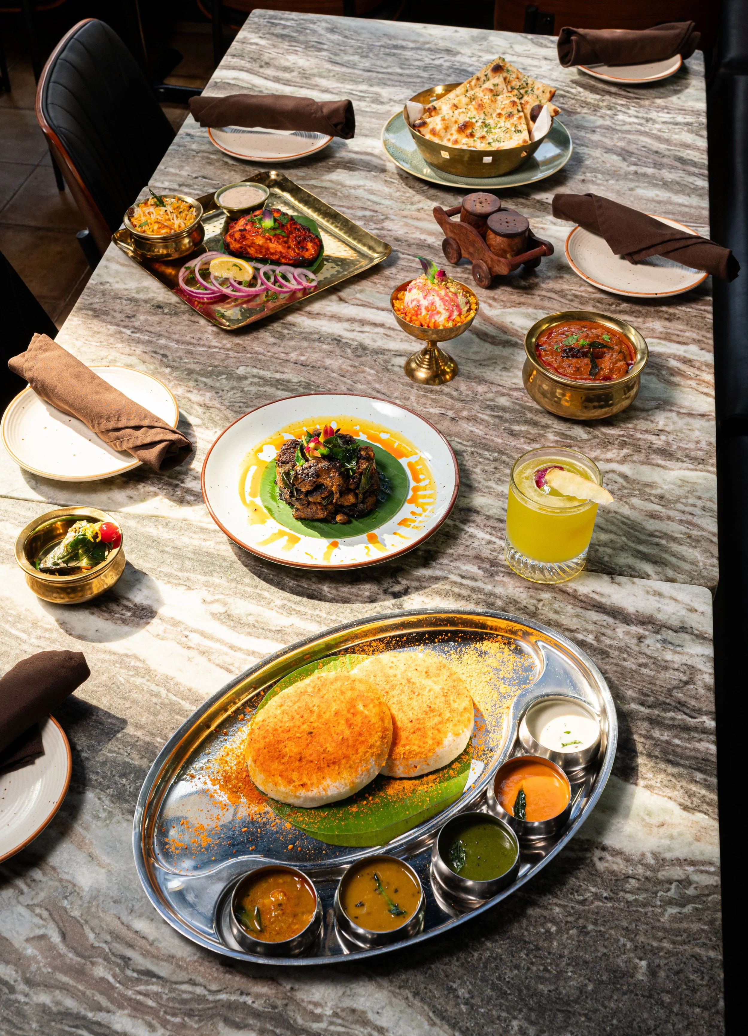 A meal from Kondattam in Calgary with naan bread, various curries, rice dish, tandoori salmon, and a yellow drink on a marble dining table.