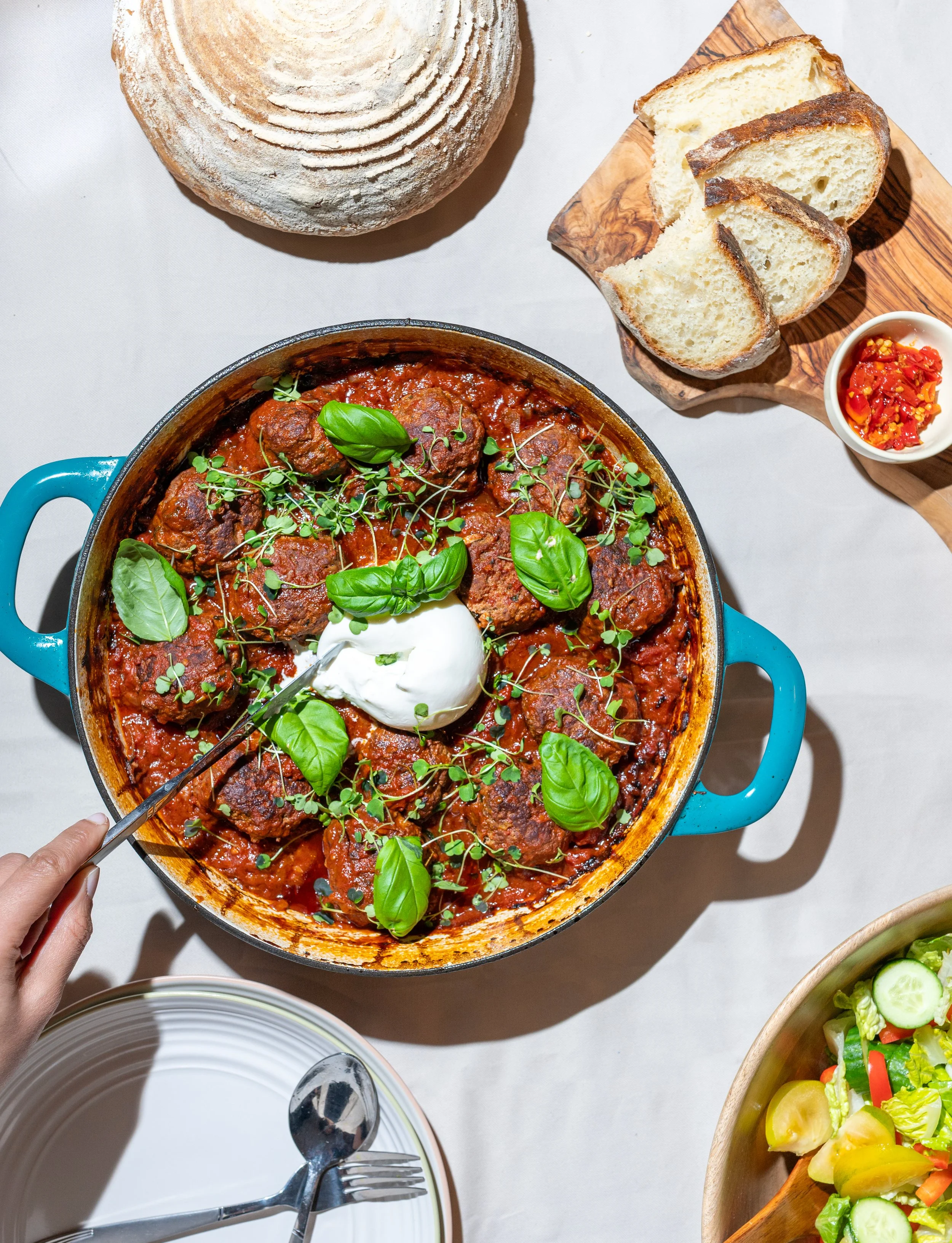 A round, blue pot containing meatballs in tomato sauce garnished with fresh basil, microgreens, and a dollop of sour cream. Surrounding items include a loaf of bread on a wooden board with slices cut, a small dish of chopped red peppers, a bowl of sa