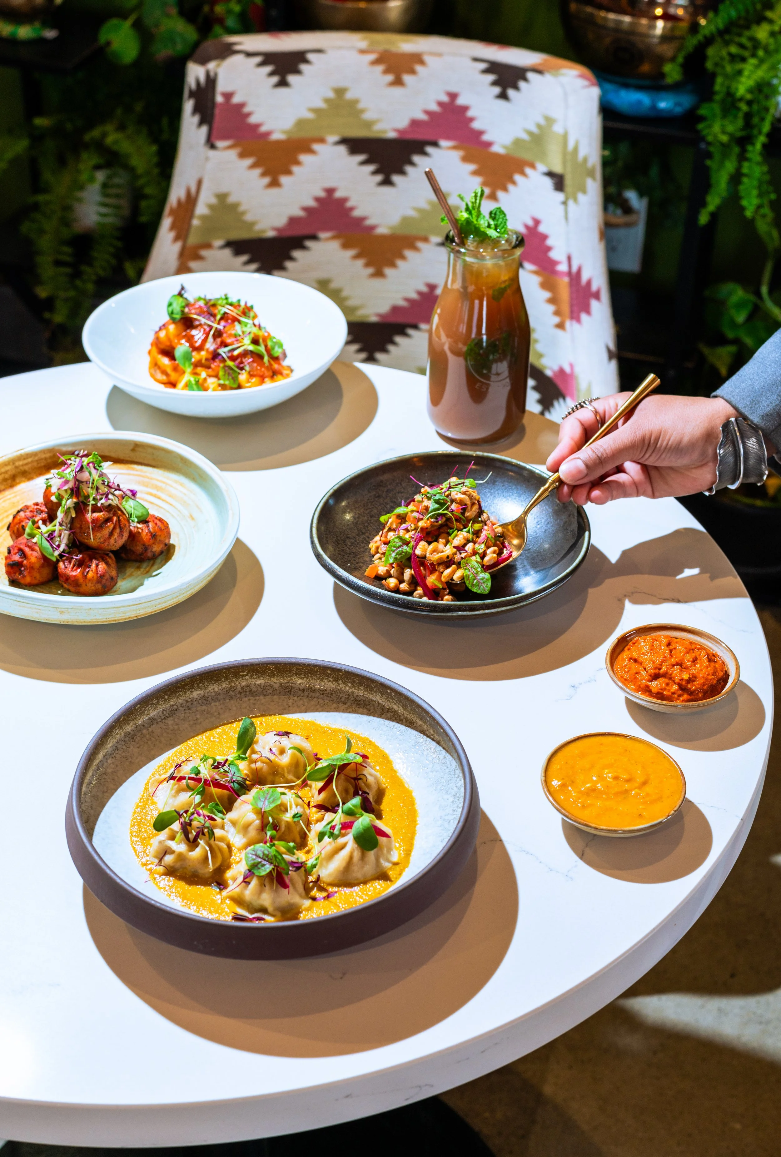 A white table with various colorful dishes from Aama Nepalese Cuisine in Cochrane, a person's hand holding a spoon, and a pink drink with mint leaves. There are patterned chairs and green plants in the background.