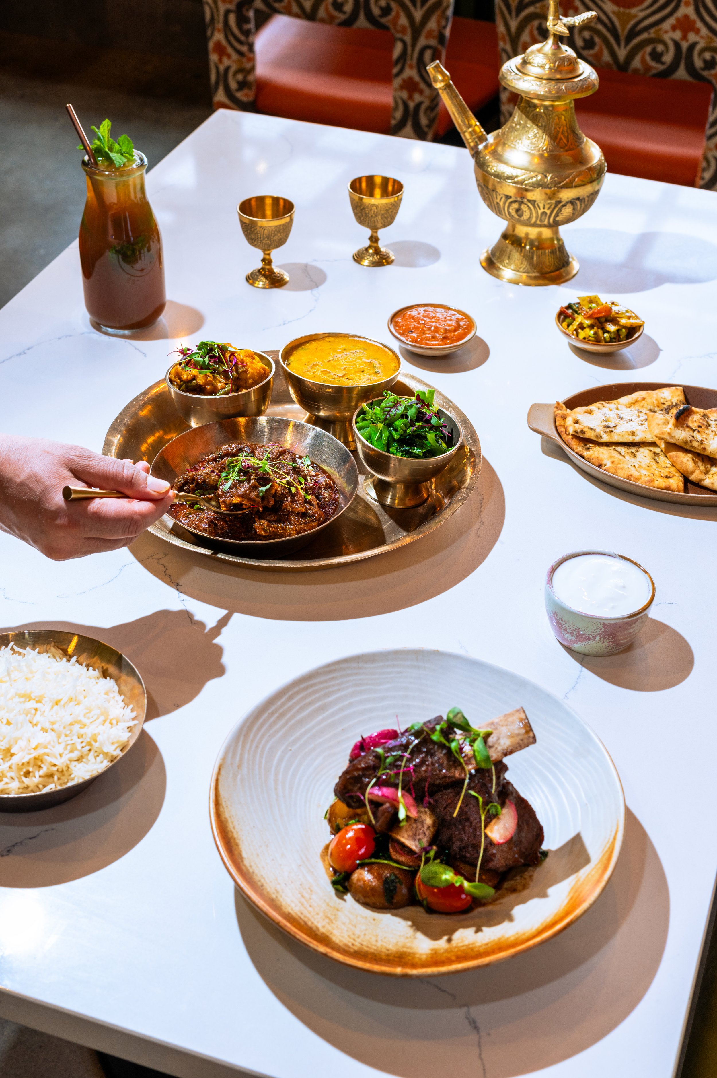 A table set with Indian cuisine, including a tray of various dishes, naan bread, rice, drinks, and utensils, with a decorative golden teapot in the background.