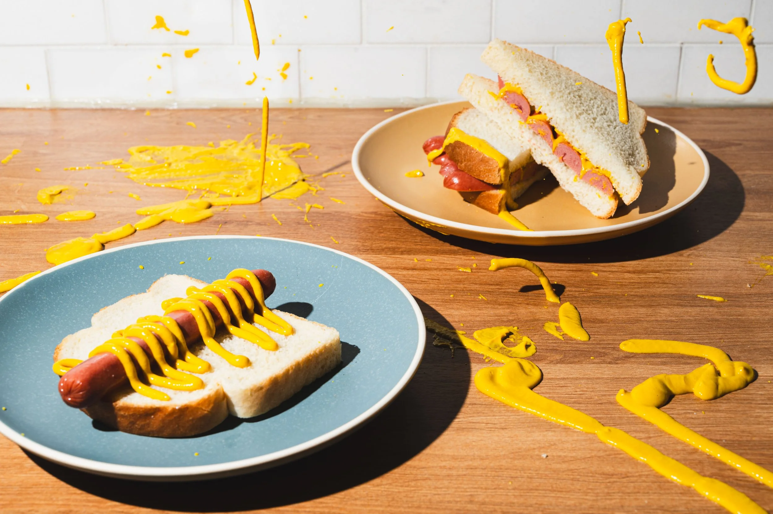 Hot dogs on blue and beige plates with mustard, with mustard splattered and dripping onto a wooden table in front of a white tiled wall.