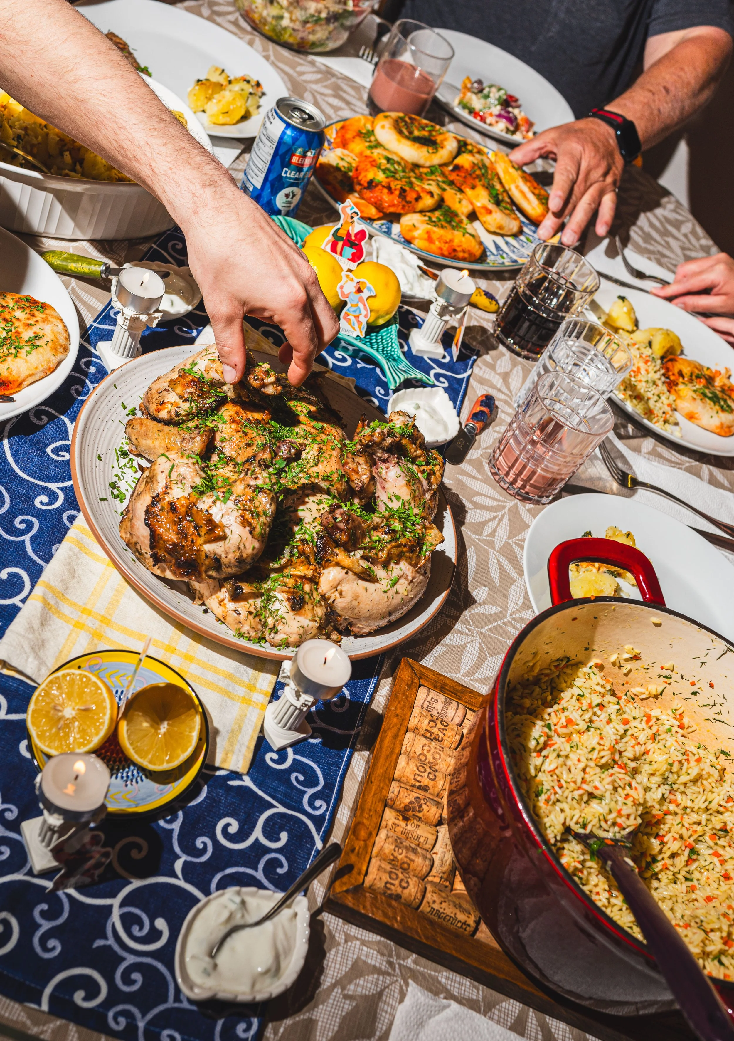 A table with a roast chicken being served, surrounded by various dishes including rice, pizza, salads, and drinks, with several people gathered around.