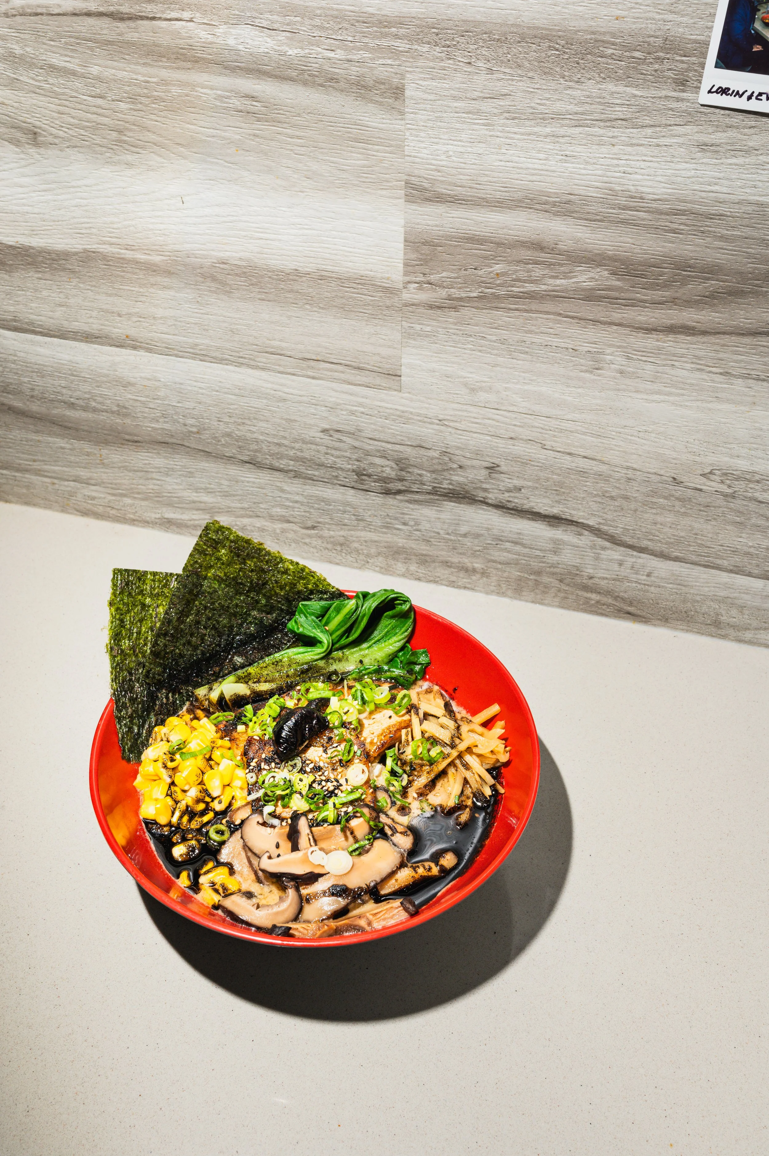 A bowl of ramen from Nan's Noodle House in Calgary with seaweed, corn, spinach, green onions, mushrooms, and slices of meat on a gray surface.