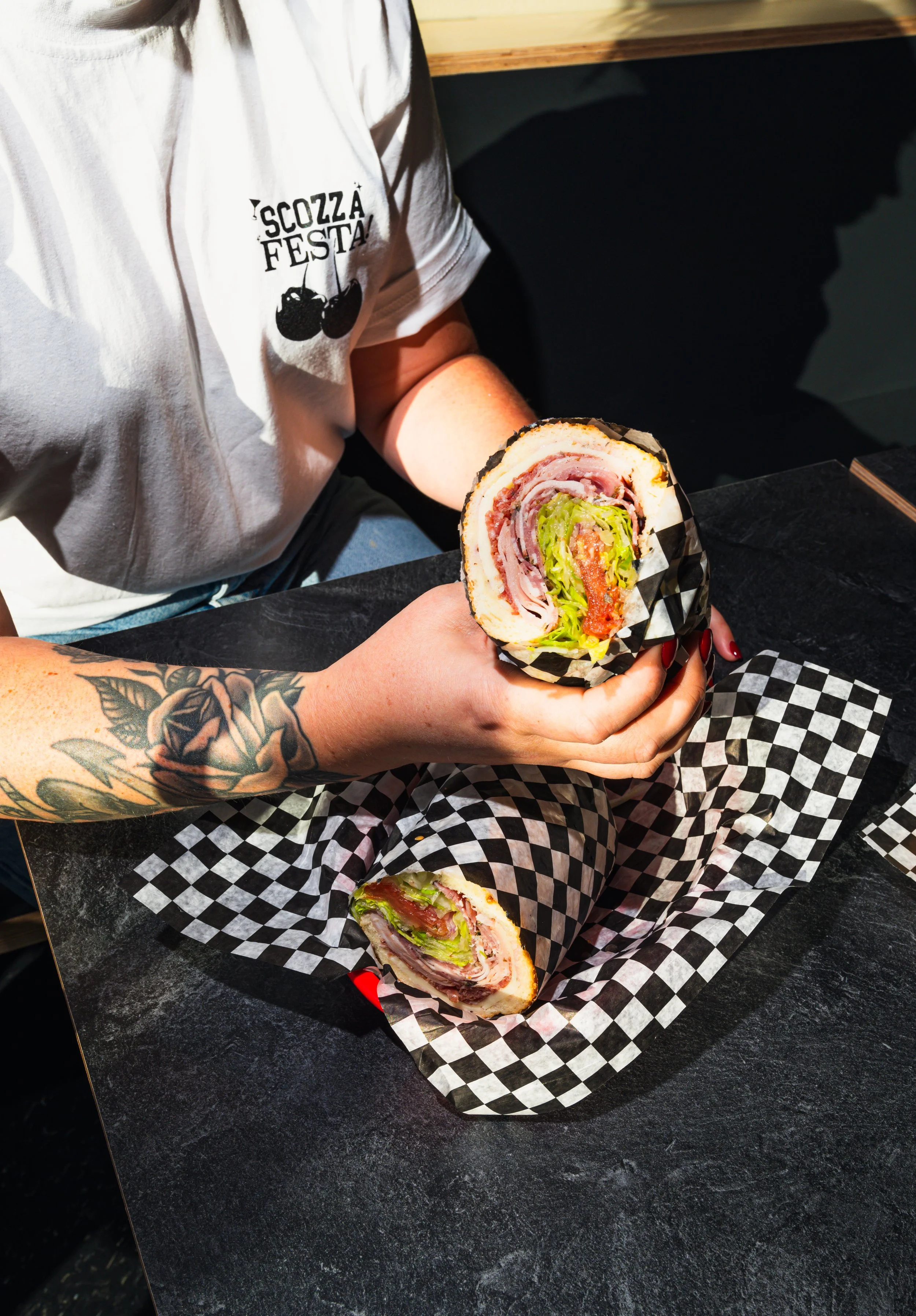 Person holding a large sandwich from Scozzafava's Deli in Calgary on black and white checkered paper.