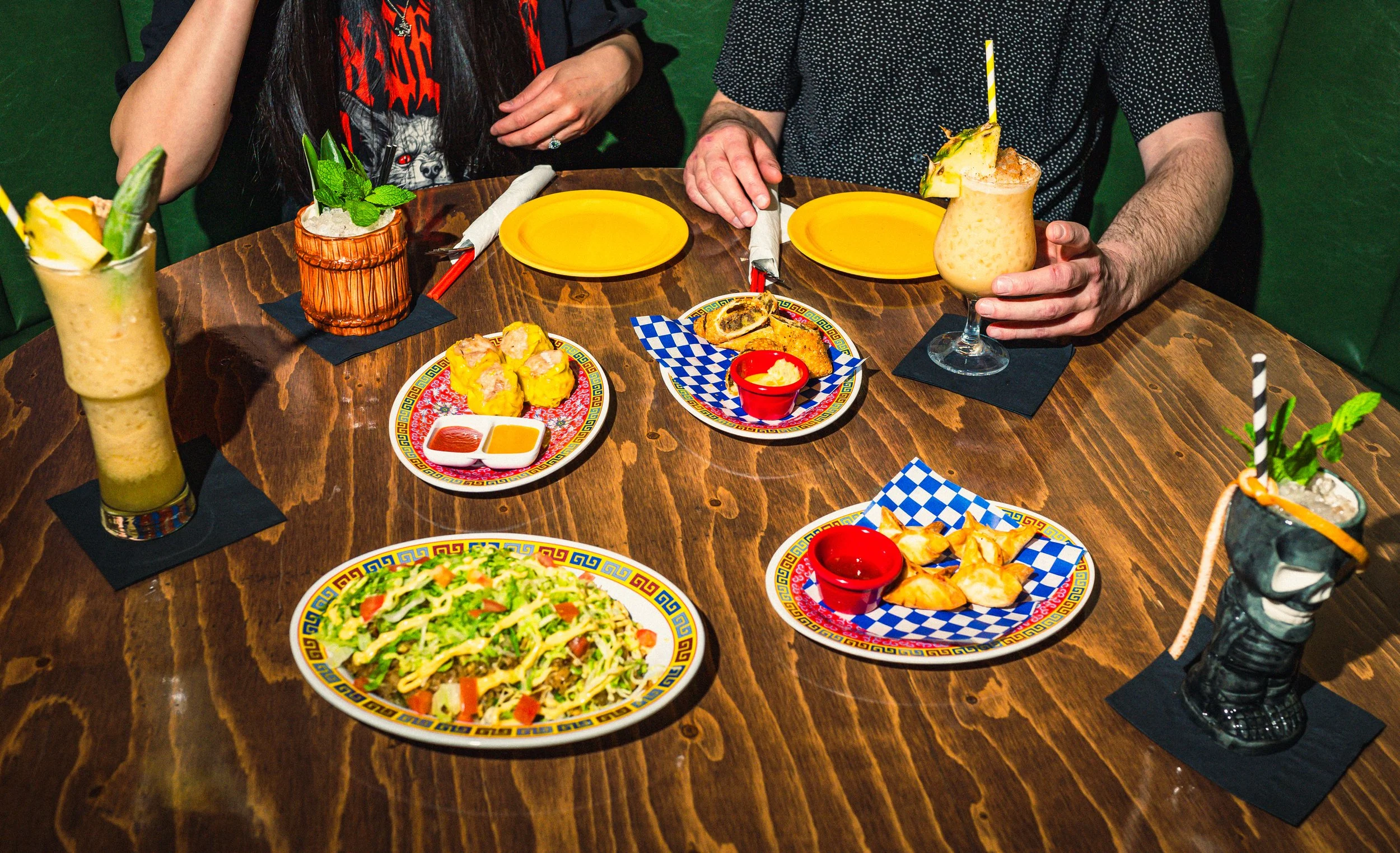 A wooden table set with dishes from Prosperity Bar in Calgary, along with colorful cocktails in a lively restaurant setting.