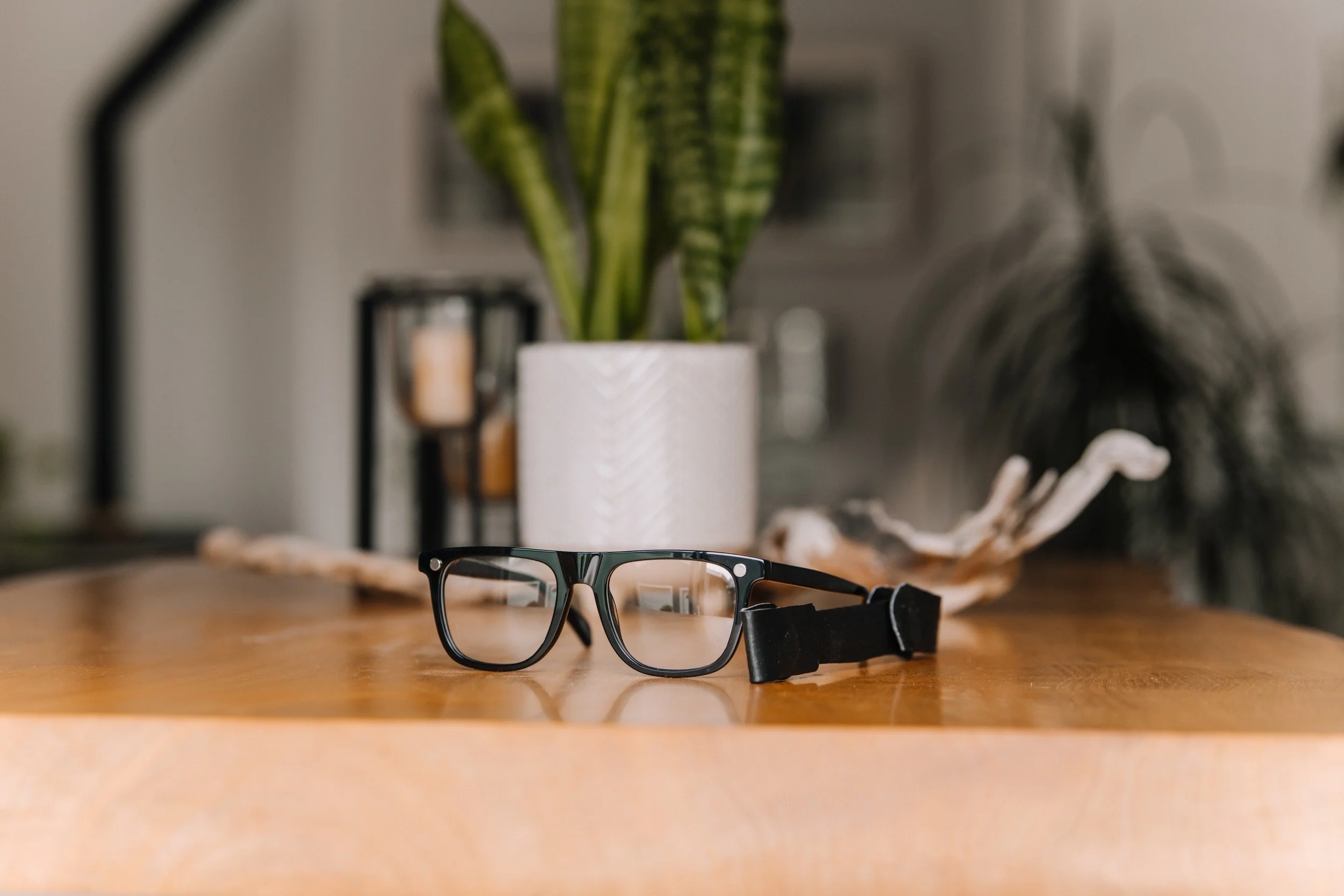 Pair of black eyeglasses with adjustable strap on a wooden table, with a potted green plant and decorative driftwood in the background.