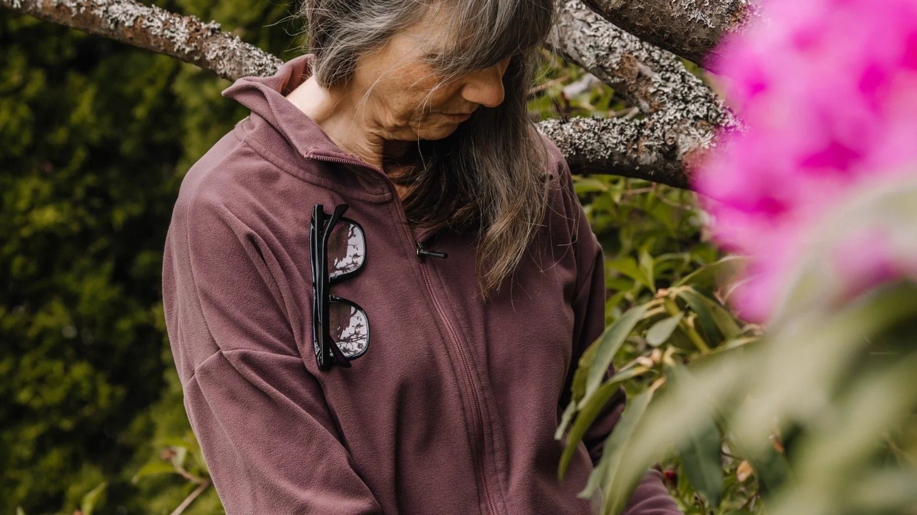 A woman with long dark hair wearing a maroon jacket with sunglasses attached, looking down at plants in a garden.