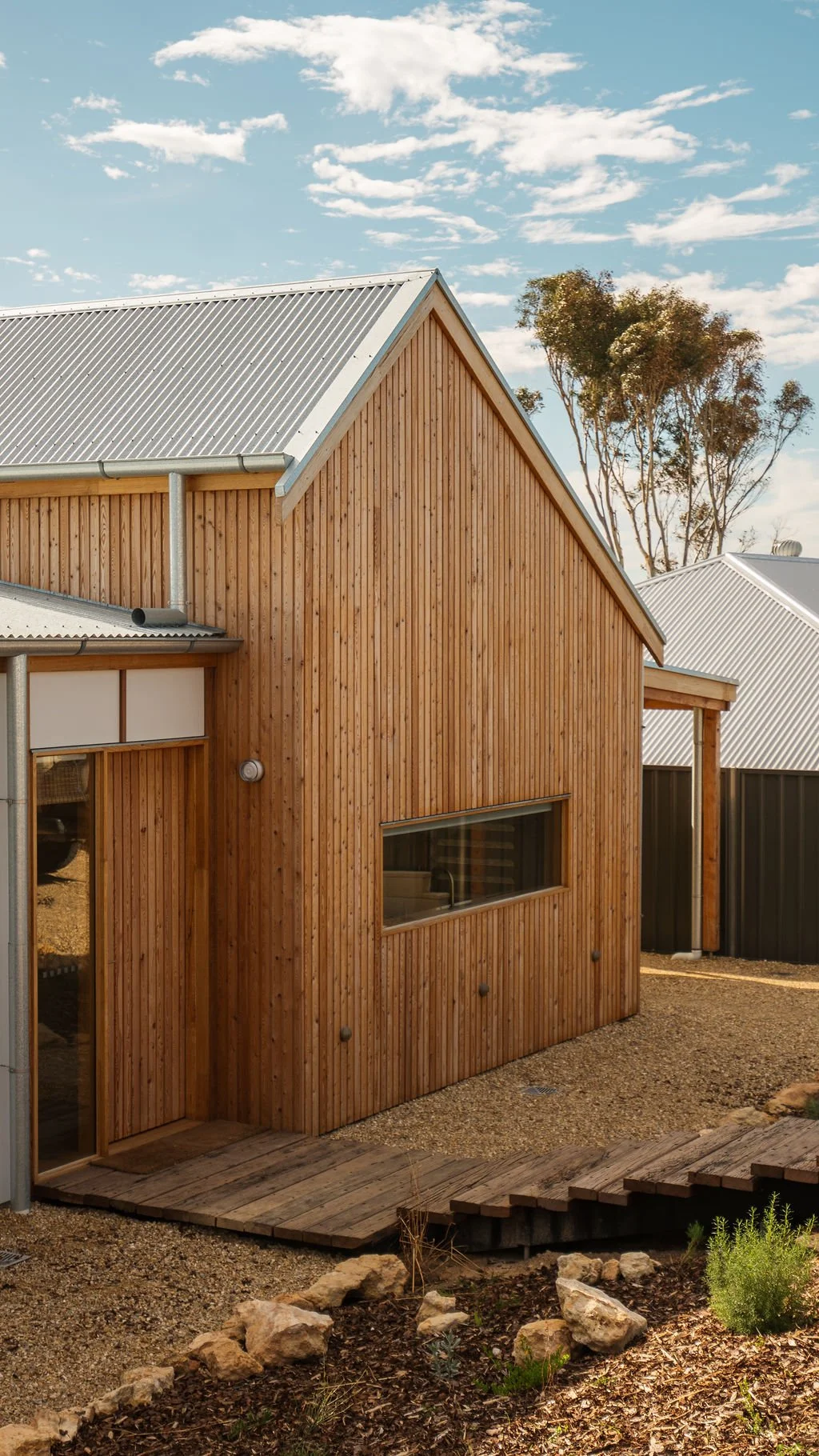 A wooden house with a metal roof, a long horizontal window, and a wooden porch in a garden with rocks and plants, under a partly cloudy sky.