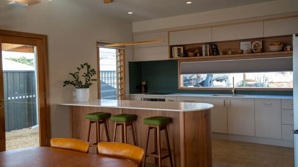 Modern kitchen with white cabinets, a wooden open shelf with dishes and books, a large window, a small bar with three green stools, and a sliding glass door leading outside.