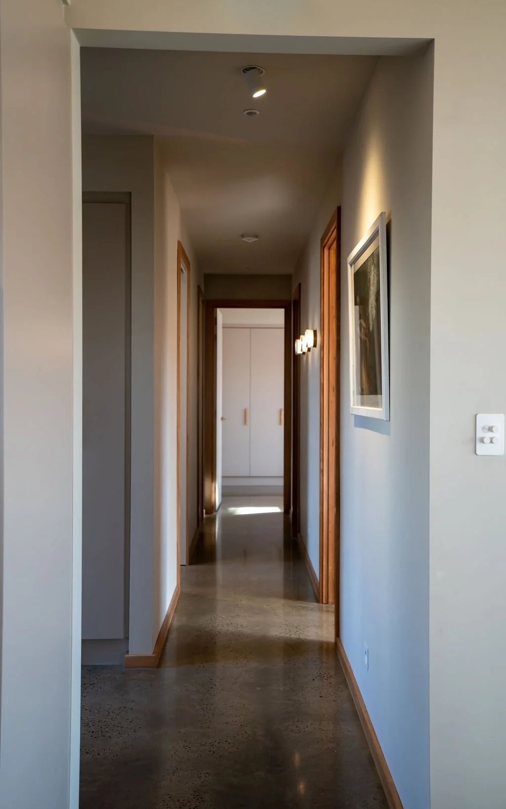 A hallway in a modern house with wood trim, white walls, framed artwork, a small wall light, and a closet at the end, illuminated by natural and ceiling light.
