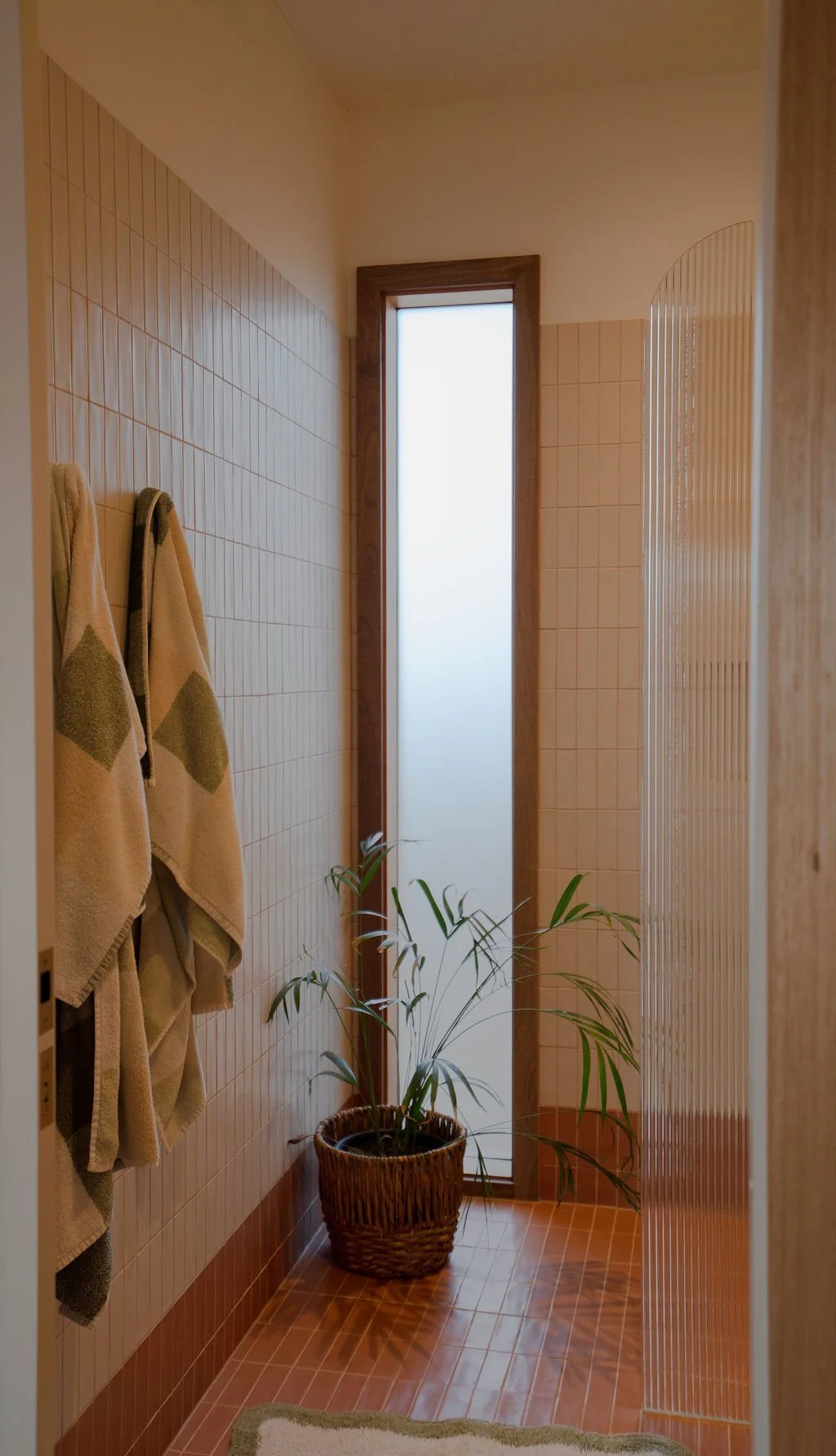 Bathroom with tiled walls and floor, dried towels hanging on hooks, potted plant in a woven basket, frosted window, and a wooden door frame.
