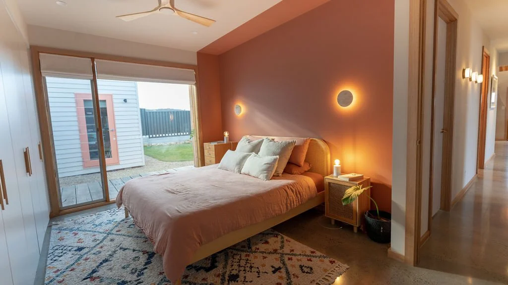 A bedroom with a bed against a pink accent wall, illuminated by wall-mounted lights and bedside lamps. The bed has white pillows and a pink blanket. There is a sliding glass door leading outside to a small deck with a view of a backyard and neighbori