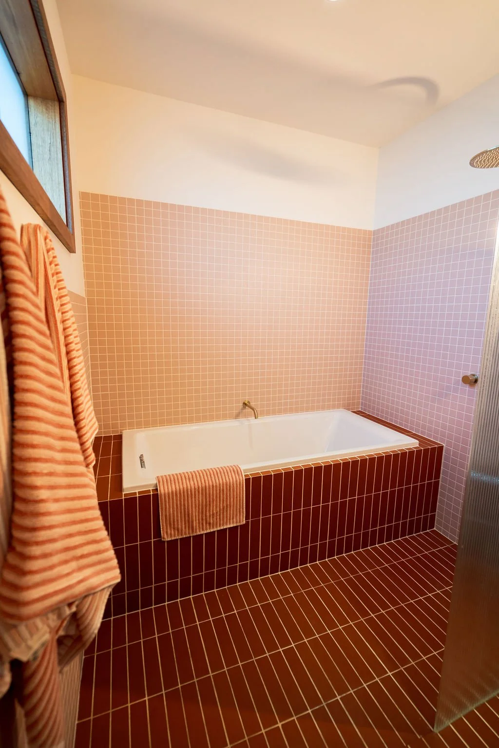 Bathroom with a white bathtub, pink tiled walls, and brown tiled floor. Pink striped towels hanging on a rack.
