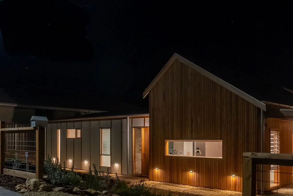 Nighttime photo of modern house with wood and white exterior, illuminated by exterior lights, showing a living space with a large window and visible interior kitchen.