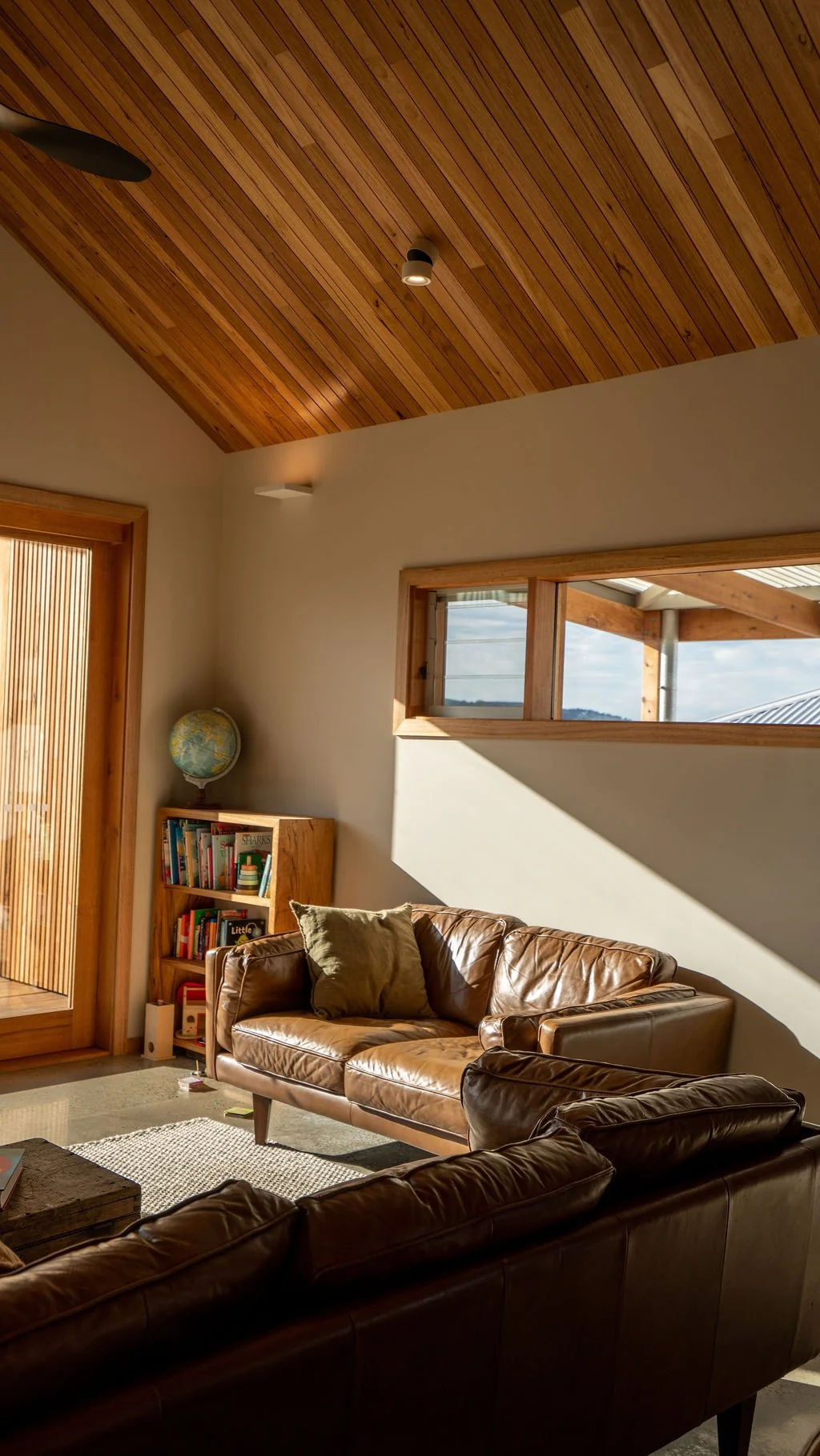 Living room with leather sofas, bookshelf with books, globe, wooden ceiling, and window letting in sunlight.
