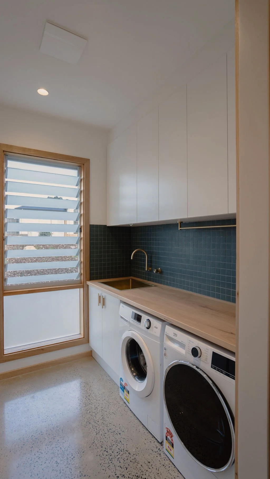 Laundry room with window, white cabinets, dark green tile backsplash, wooden countertop, gold faucet, washing machine, and dryer.