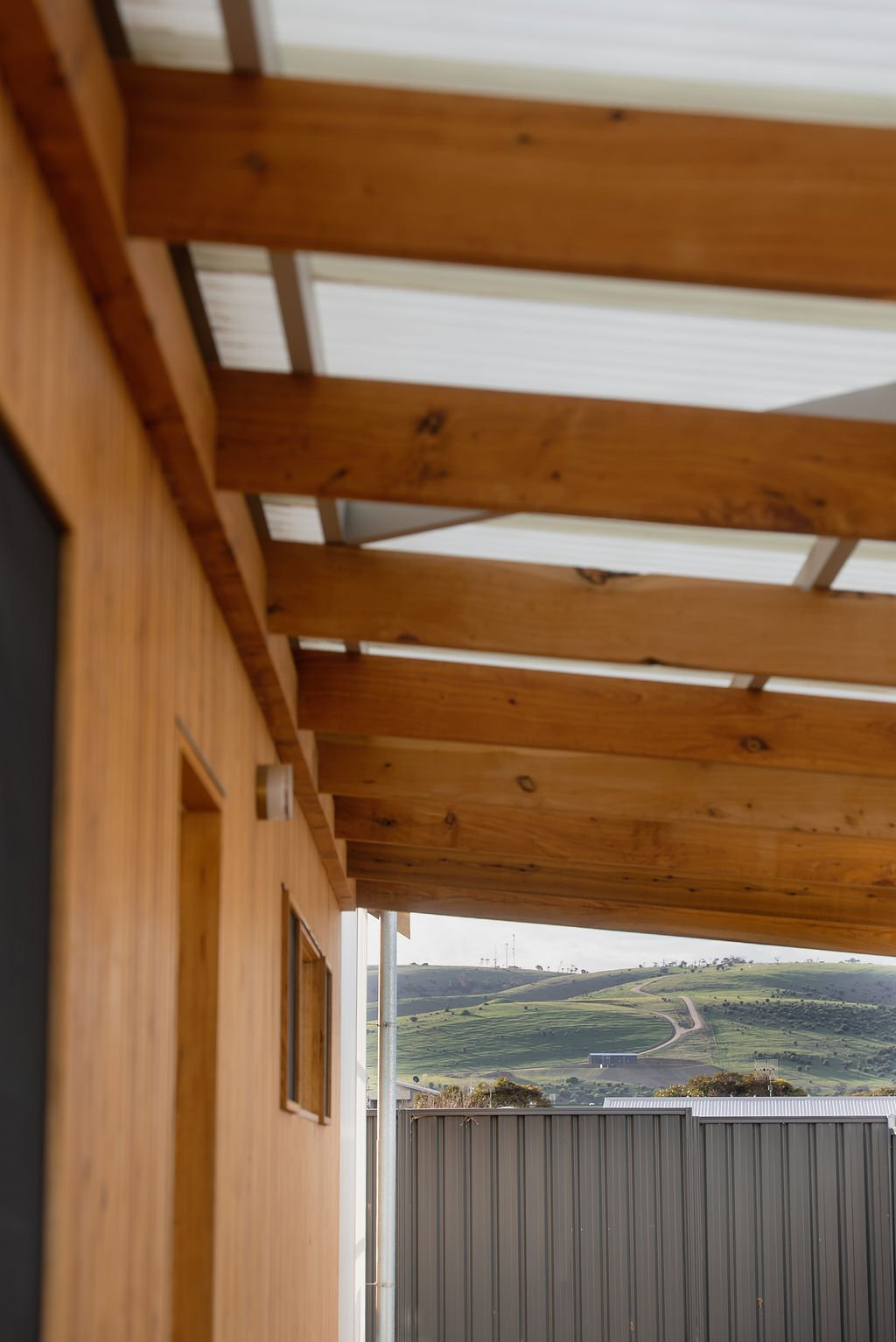 View of a hillside landscape with green rolling hills and winding roads, seen from a wooden porch or balcony with a roof supported by wooden beams.